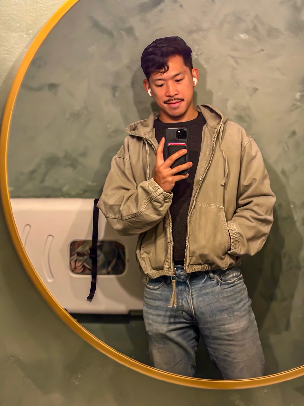 A man takes a mirror selfie in a bathroom. He is wearing a jacket and jeans.