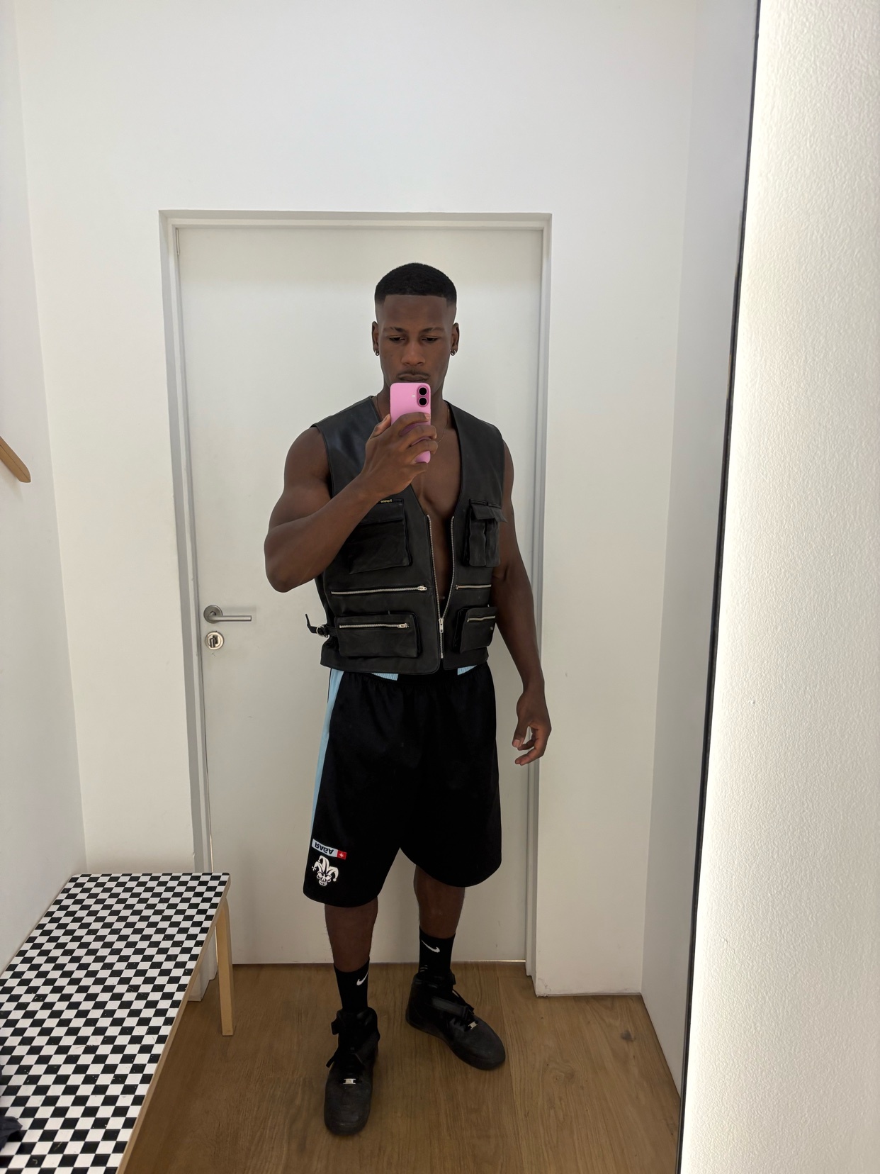 A man takes a mirror selfie, showcasing his physique and fashion choices. He is shirtless, wearing a black vest and shorts.