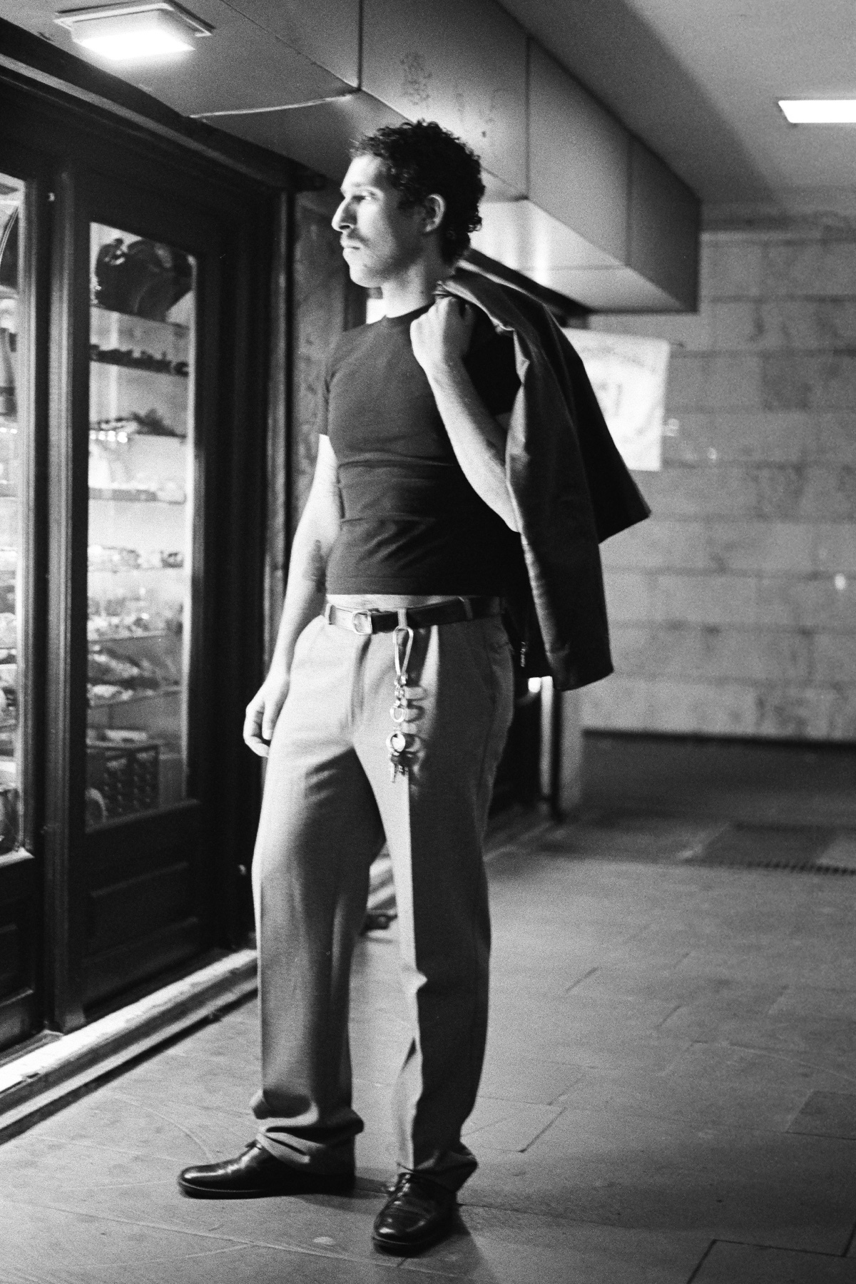 A man in a black t-shirt and suit jacket poses in front of a shop window. The black and white photo captures a moment of urban style.