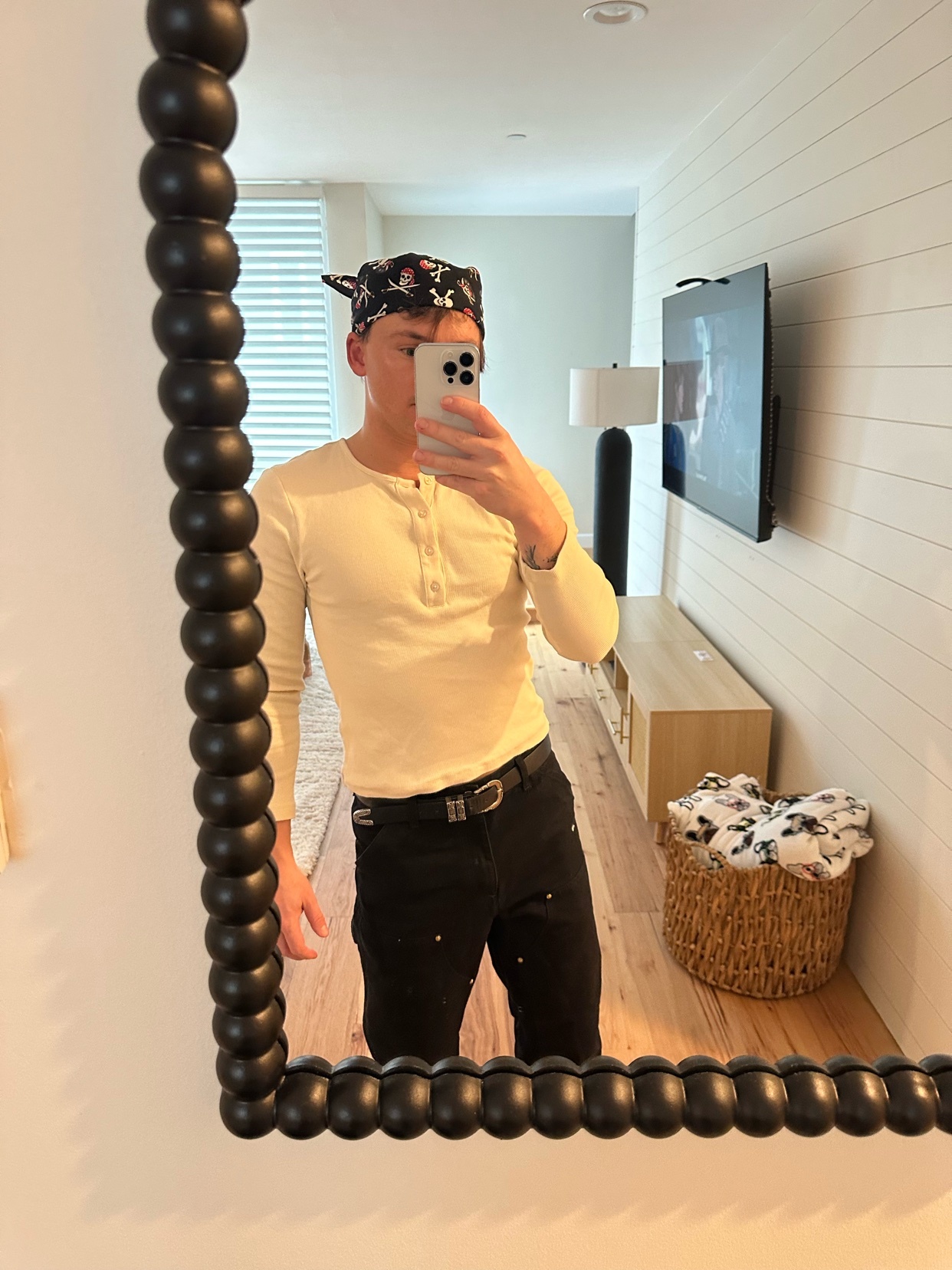 A man takes a mirror selfie, showcasing his outfit. He wears a bandana, long-sleeved shirt, and black pants.