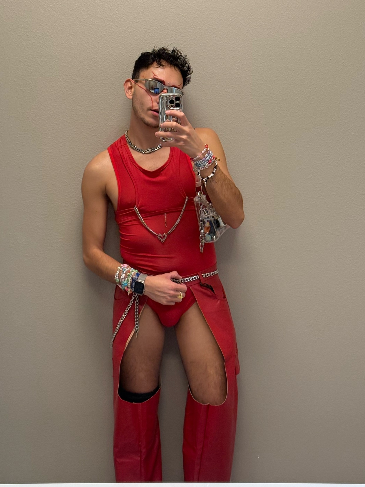 A man takes a mirror selfie, showcasing a bold red outfit. The outfit includes a tank top, chaps, and accessories.