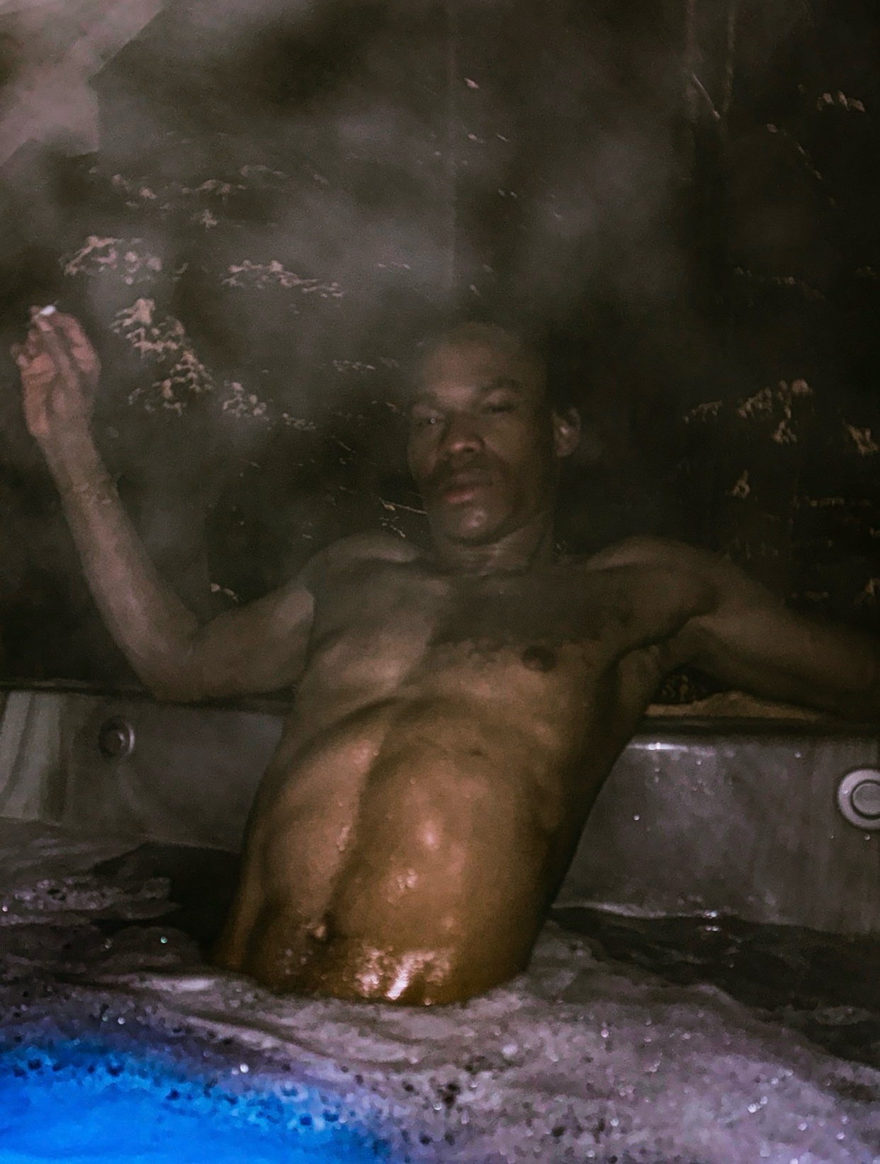 A shirtless man relaxes in a hot tub, steam rising around him. The photo is taken from a low angle, emphasizing the man's physique.