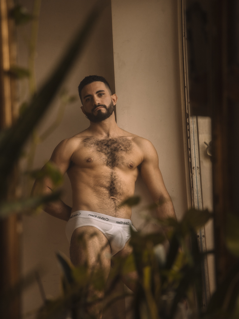 A man poses shirtless, wearing white underwear. The photo is framed by plants, creating a natural and intimate setting.