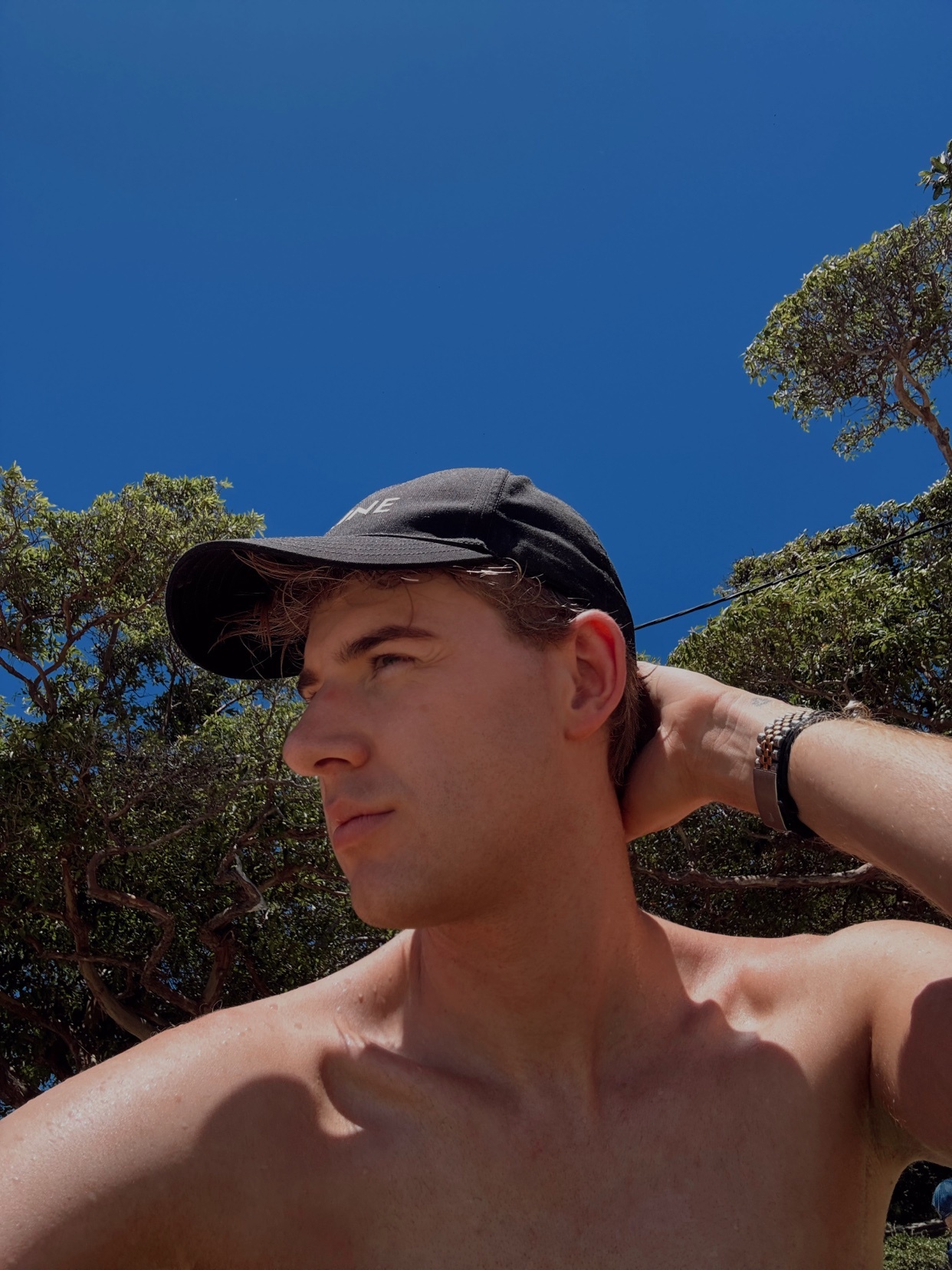 A young man wearing a black baseball cap and no shirt poses outdoors under a bright blue sky. He is looking off to the side with his hand on the back of his head, with trees visible in the background.