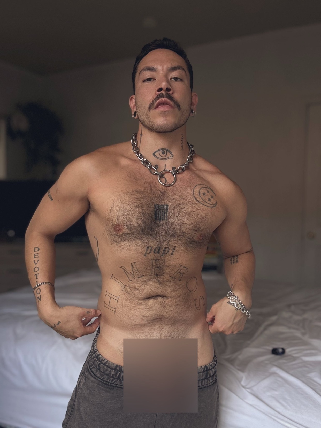 A man with tattoos and a mustache poses shirtless in front of a bed, wearing low-rise pants.