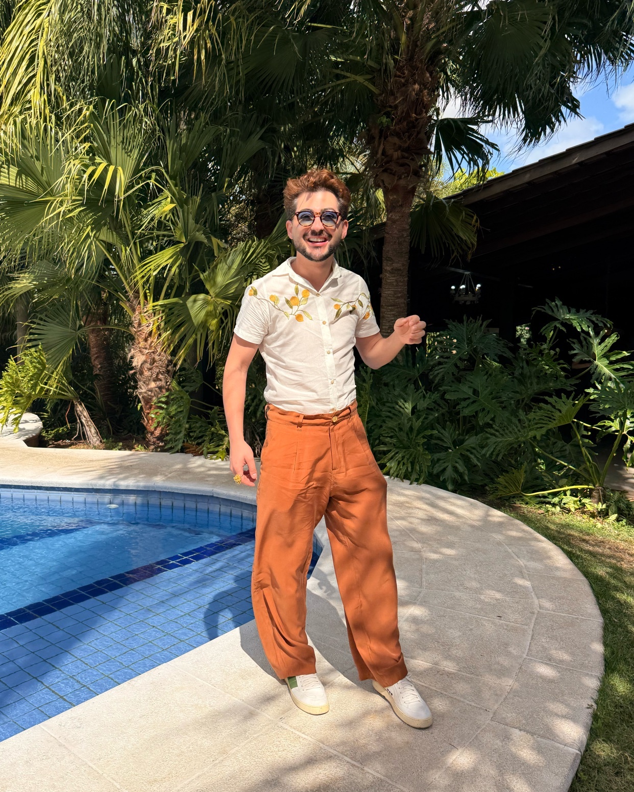 A man in stylish resort wear poses poolside with lush greenery and palm trees in the background. He wears a short-sleeved button-down shirt and wide-leg orange pants, accessorized with sunglasses.