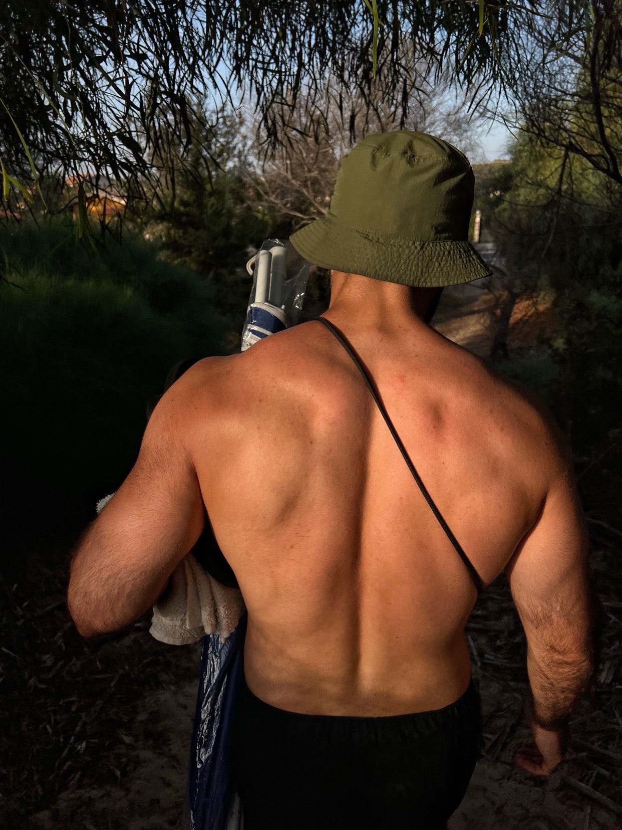 A man with a muscular back walks away from the camera, shirtless and wearing a bucket hat. He is carrying items over his shoulder.