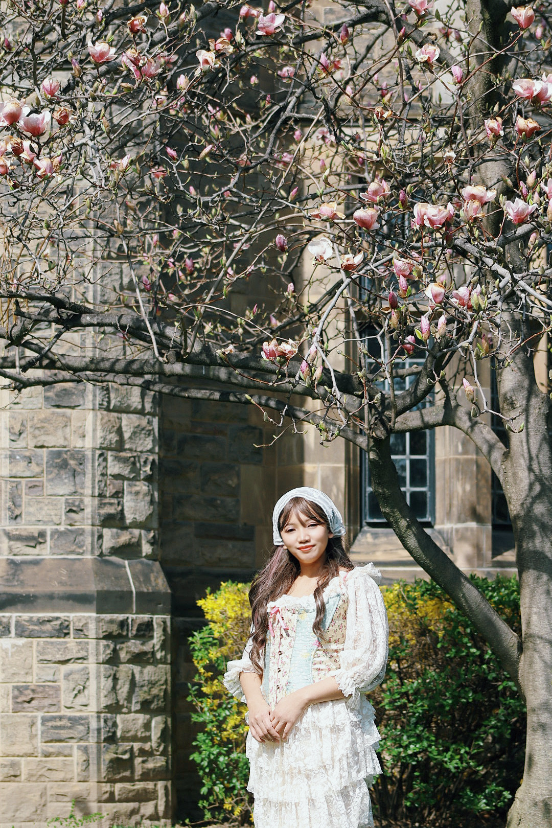 A young woman in a white lace dress stands in front of a stone building, with a blooming magnolia tree providing a soft pink canopy overhead. The scene evokes a sense of spring and vintage charm.