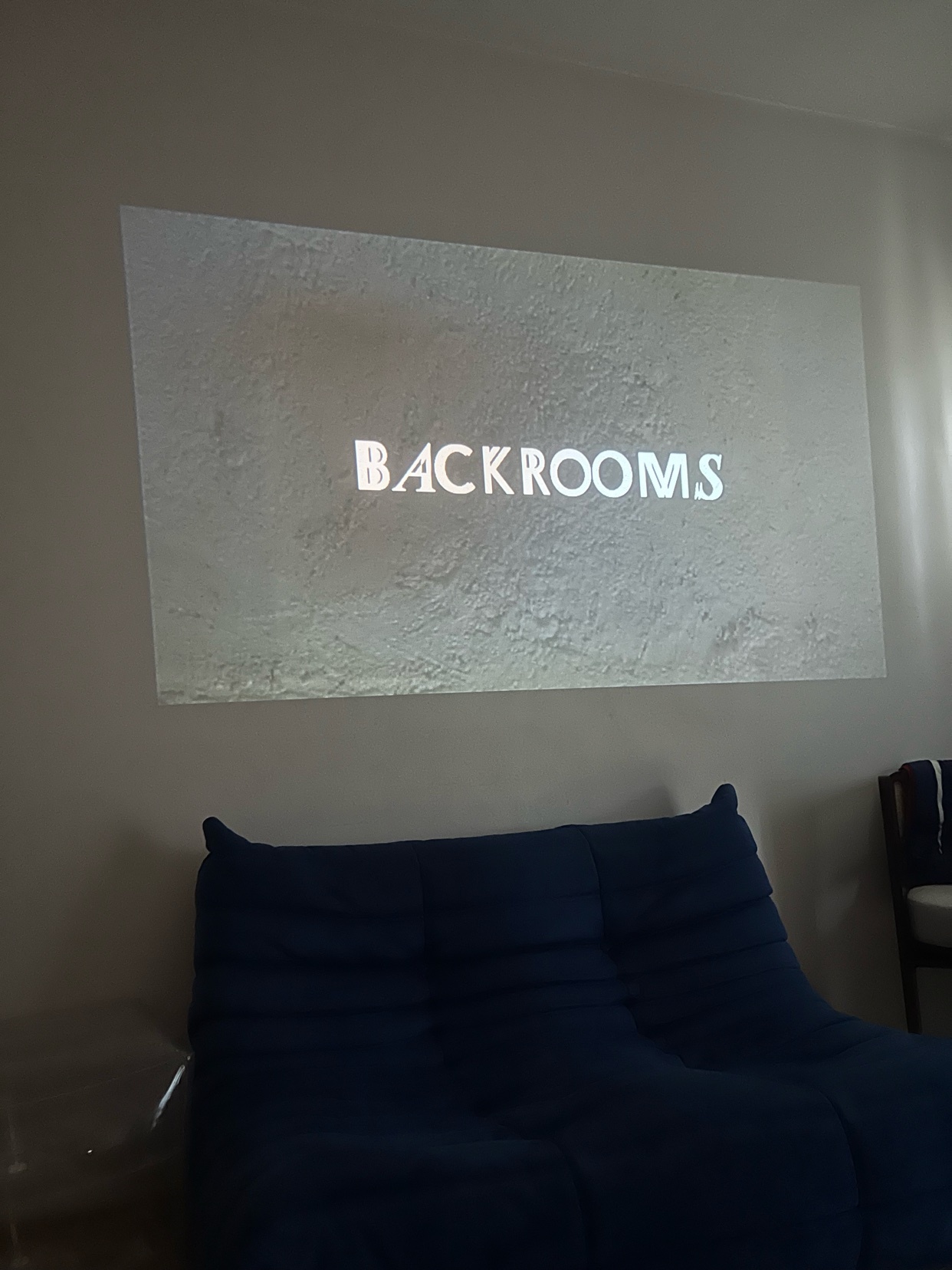 A projector displays the word "BACKROOMS" onto a textured wall above a dark blue couch.