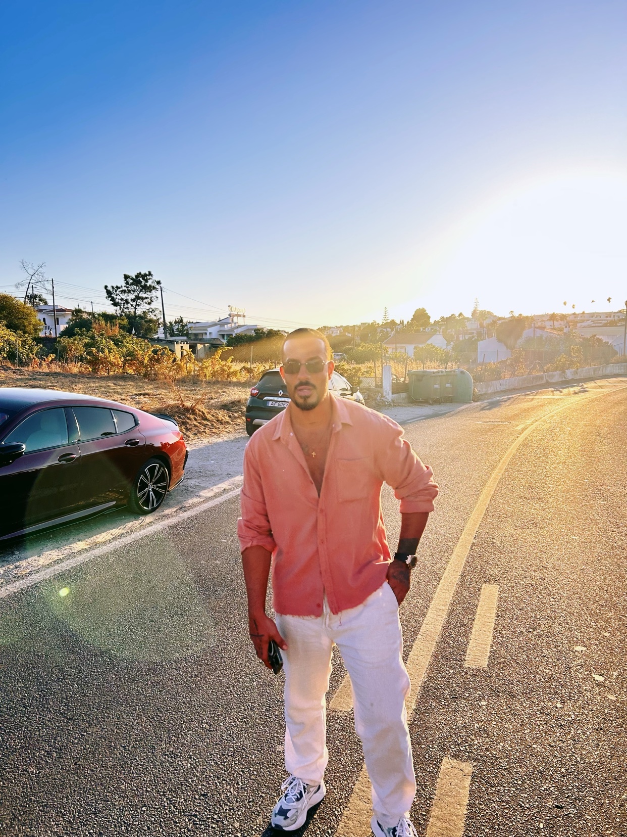 A man stands on a road during golden hour, wearing a pink button-down shirt, white pants, and sunglasses. A car is parked to his left, and the sun sets in the background.