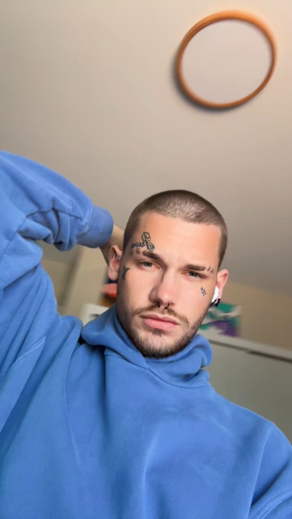 A man poses for a selfie, wearing a blue hoodie. He has tattoos on his face and a short haircut.