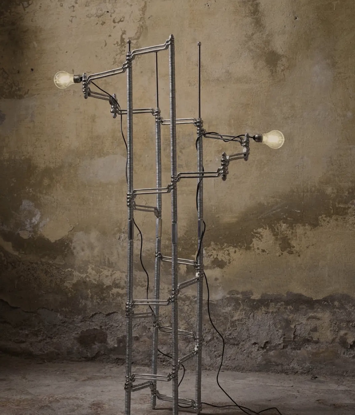An artistic lamp made of metal with a unique design. The lamp is placed in front of a textured wall.