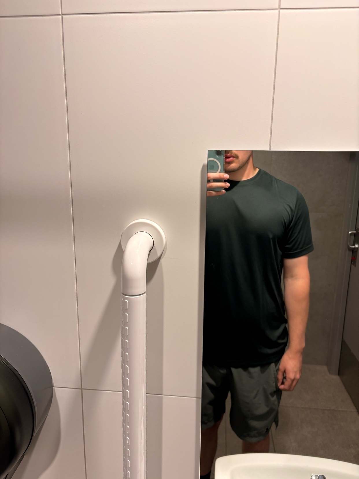 A man takes a mirror selfie in a bathroom. He is wearing a t-shirt and shorts.