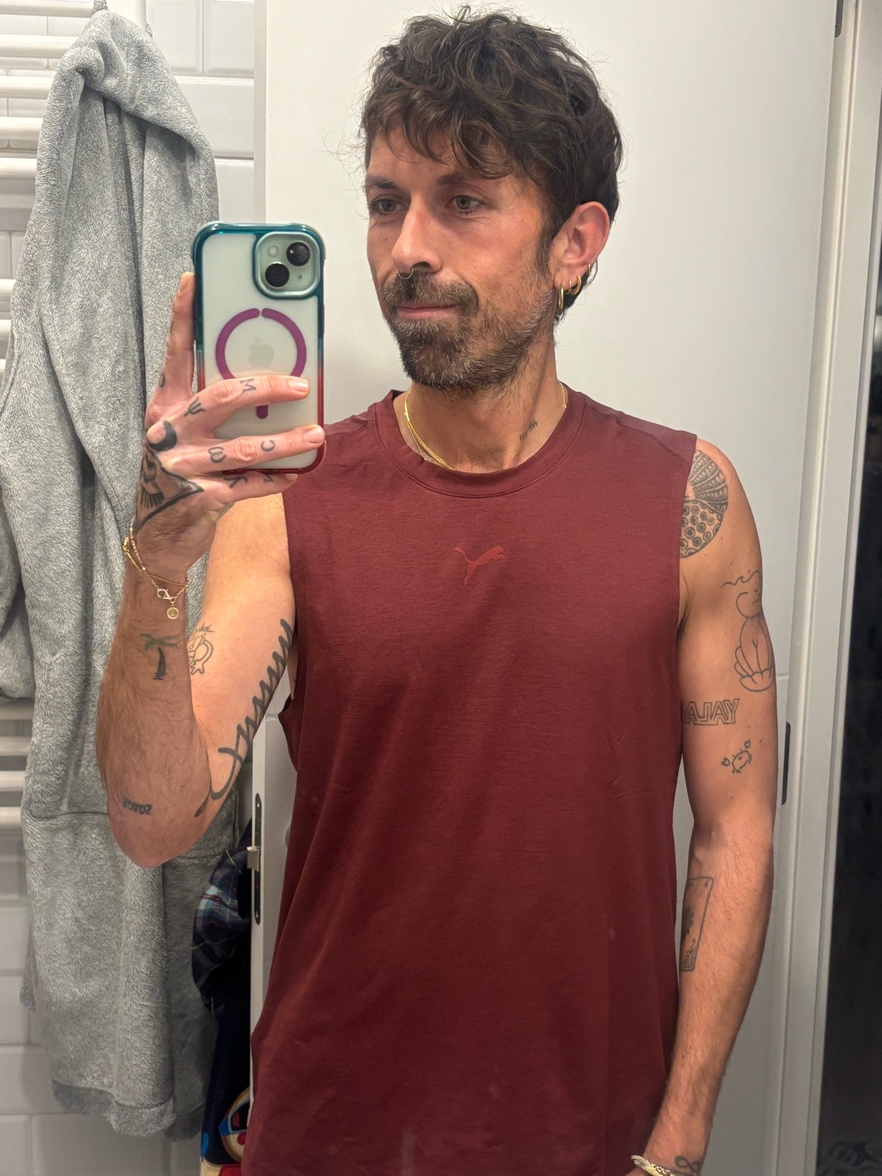 A man with short, dark, wavy hair and stubble takes a selfie in a bathroom. He is wearing a maroon sleeveless shirt and has tattoos on his arms.