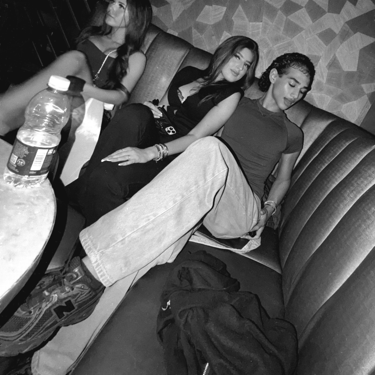 Three young women are seated on a plush couch in a dimly lit setting, possibly a club or lounge. One woman in the foreground wears light-wash jeans and a dark top, while the other two are dressed in darker attire.