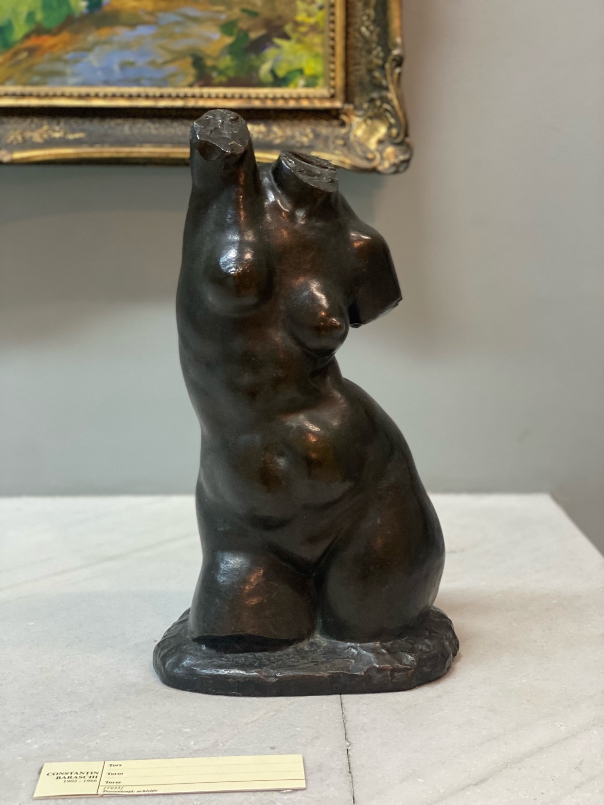A bronze sculpture of a female torso sits on a pedestal in front of a framed painting. The sculpture is dark brown and appears to be in a classical style, with the head and limbs missing.