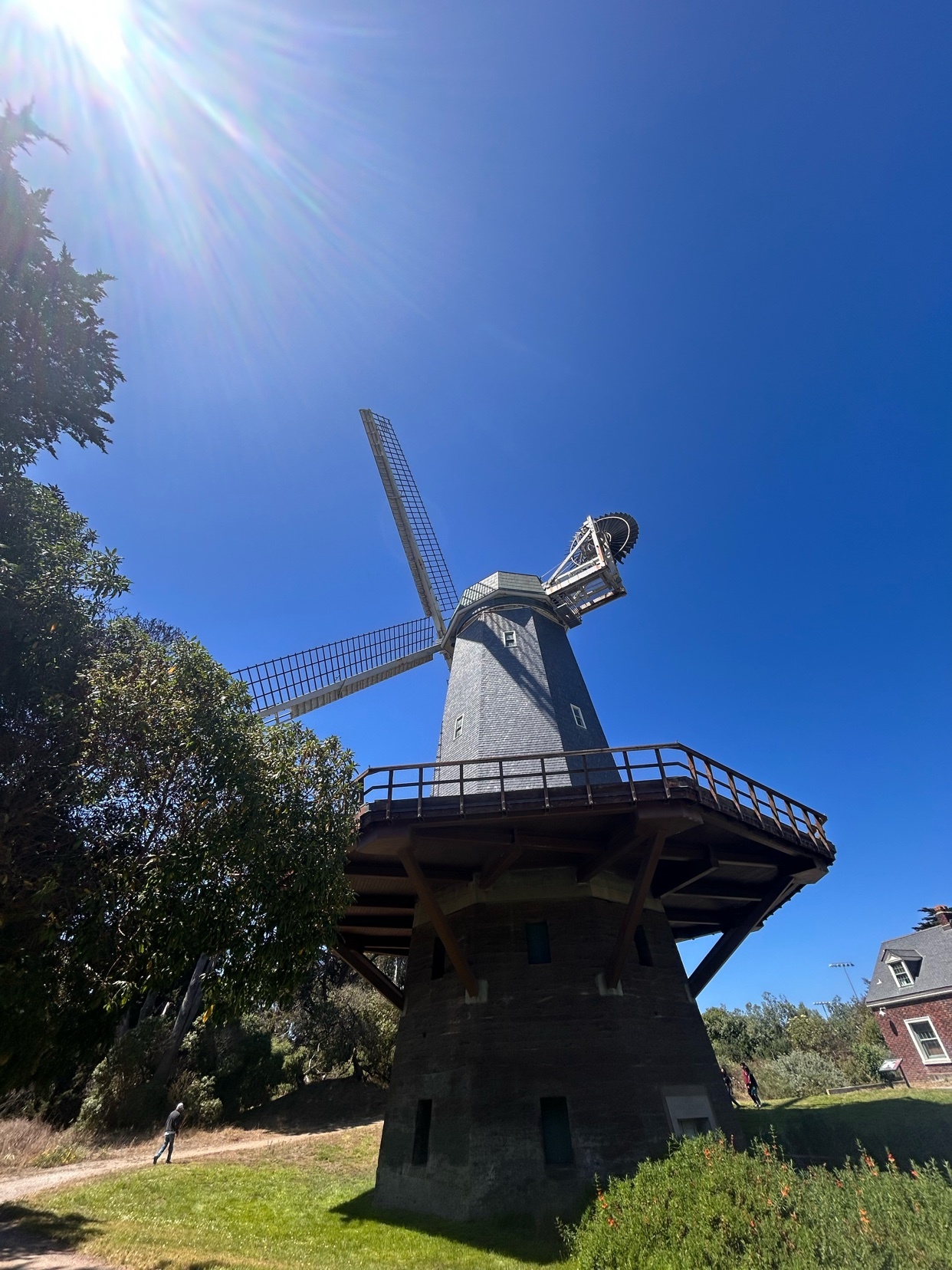 A Dutch windmill stands tall against a clear blue sky, with the sun shining brightly and casting lens flare. A person is visible walking on a path near the base of the windmill.