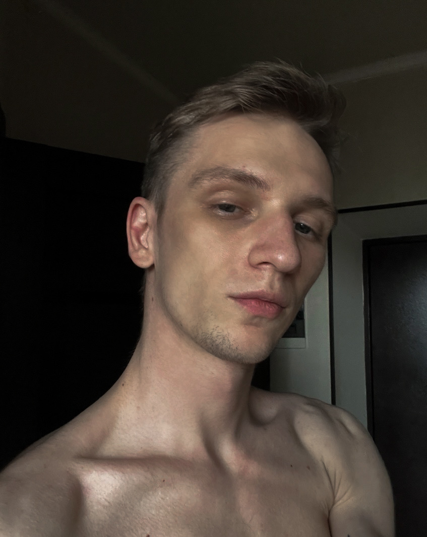 A close-up selfie of a shirtless man with blond hair and light skin. He is looking directly at the camera with a neutral expression. Cat ears are visible on his head.