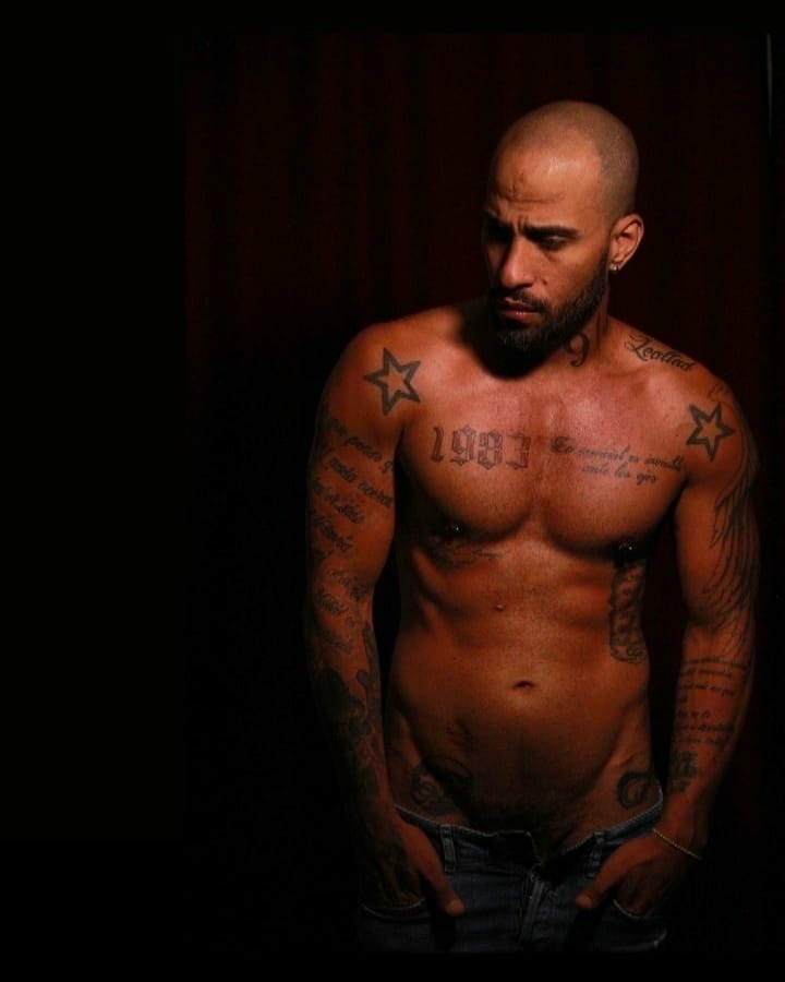 A shirtless, bald man with a beard and tattoos poses with his hands in his jeans. He has piercings on his chest and a star tattoo on his shoulder.