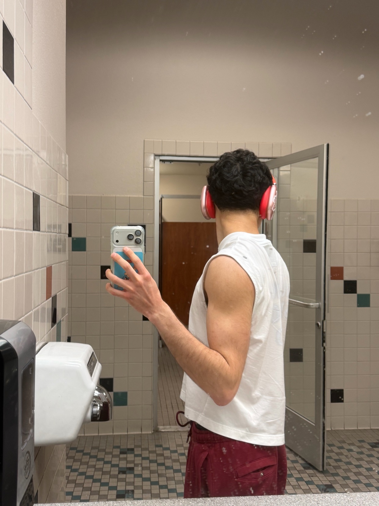 A man takes a mirror selfie in a gym bathroom, wearing a white sleeveless shirt and red shorts, with red headphones on.