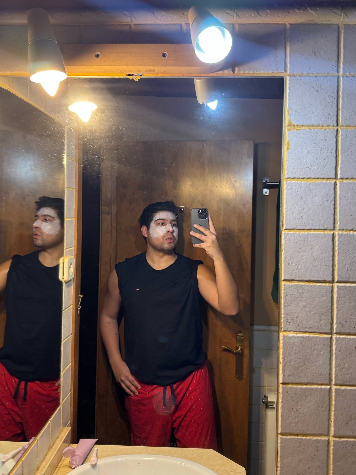A man takes a selfie in the mirror while wearing a white face mask and a black tank top. He is also wearing red shorts and is standing in a bathroom.