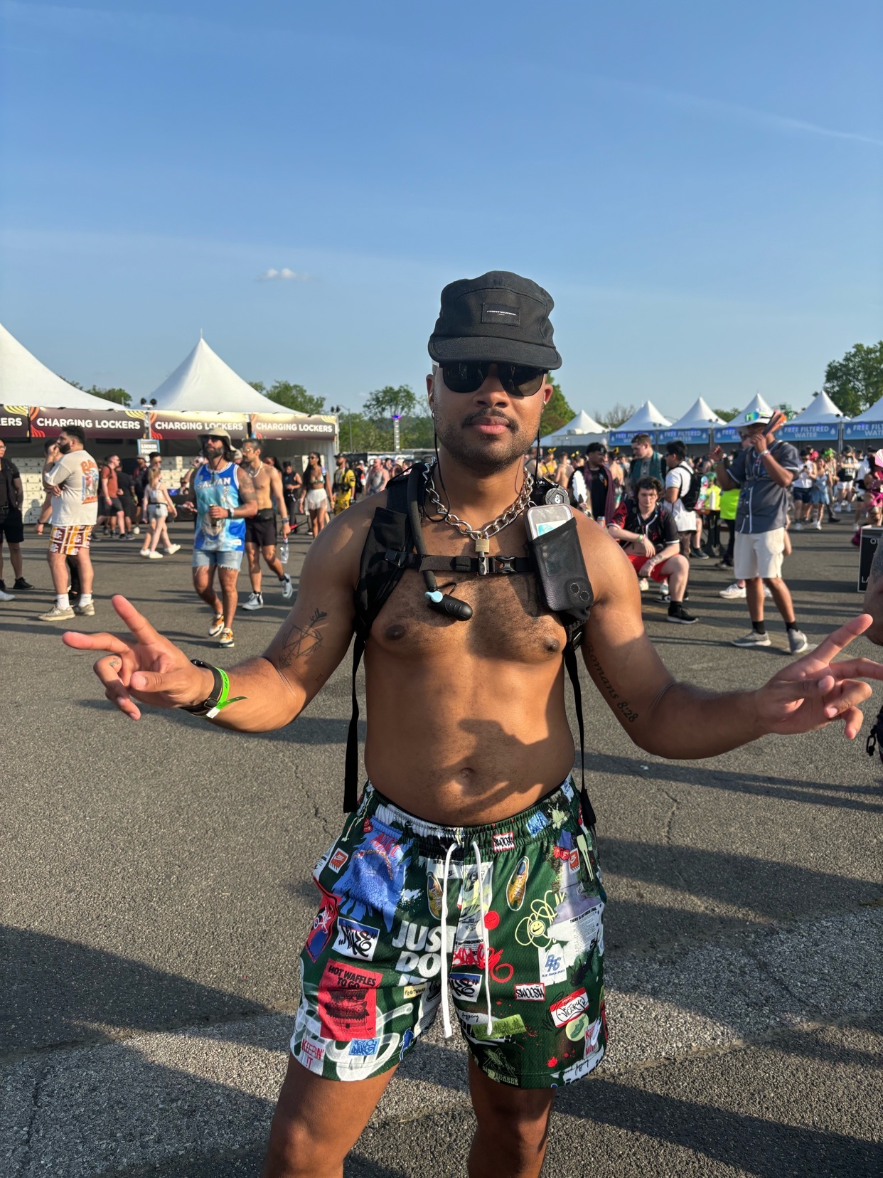 A man poses shirtless at an outdoor festival. He wears shorts, a hat, and sunglasses.
