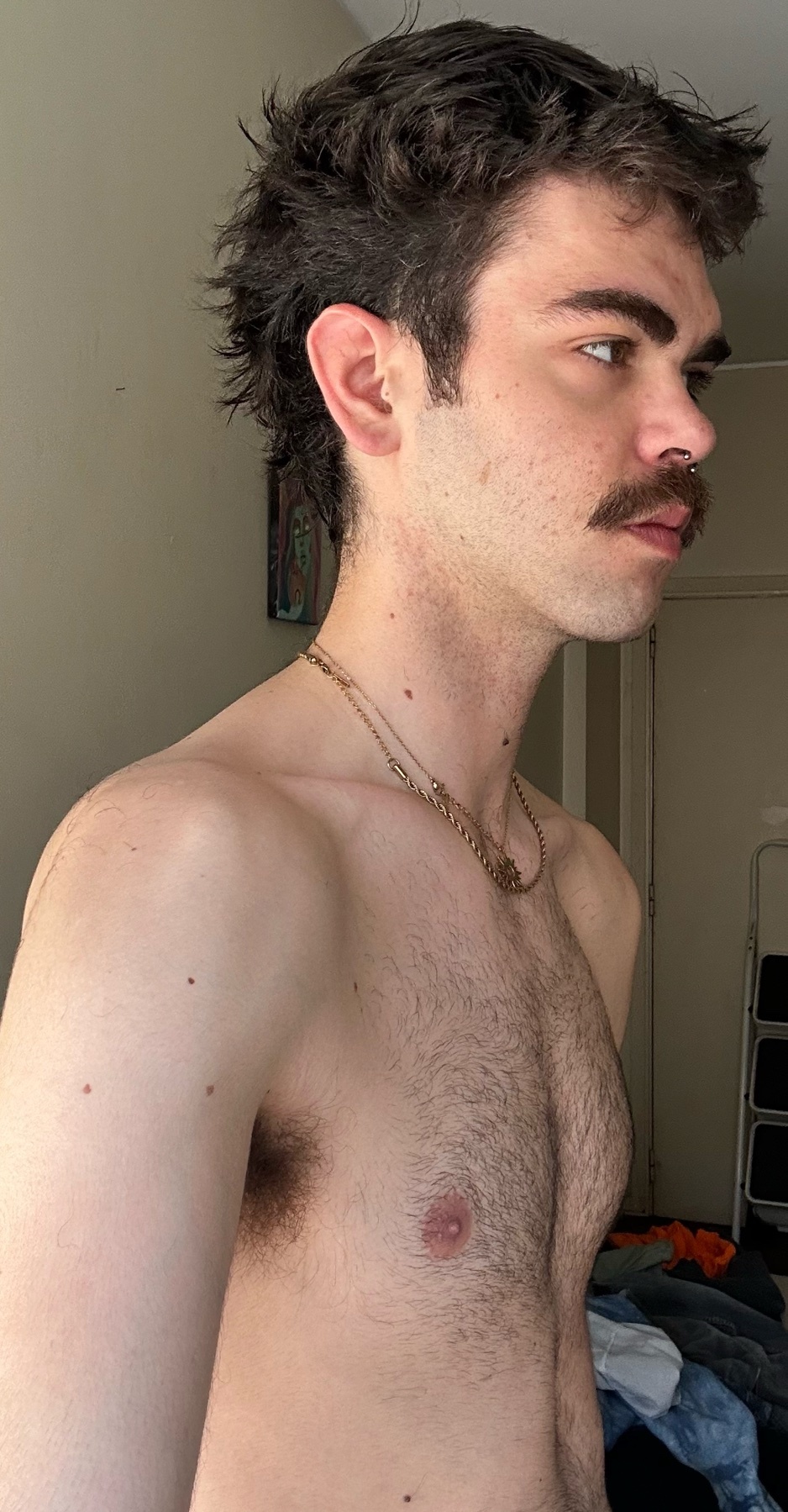 A man with a mustache and dark, messy hair is shown in profile, looking to the right. He is wearing a necklace and has visible chest and armpit hair.