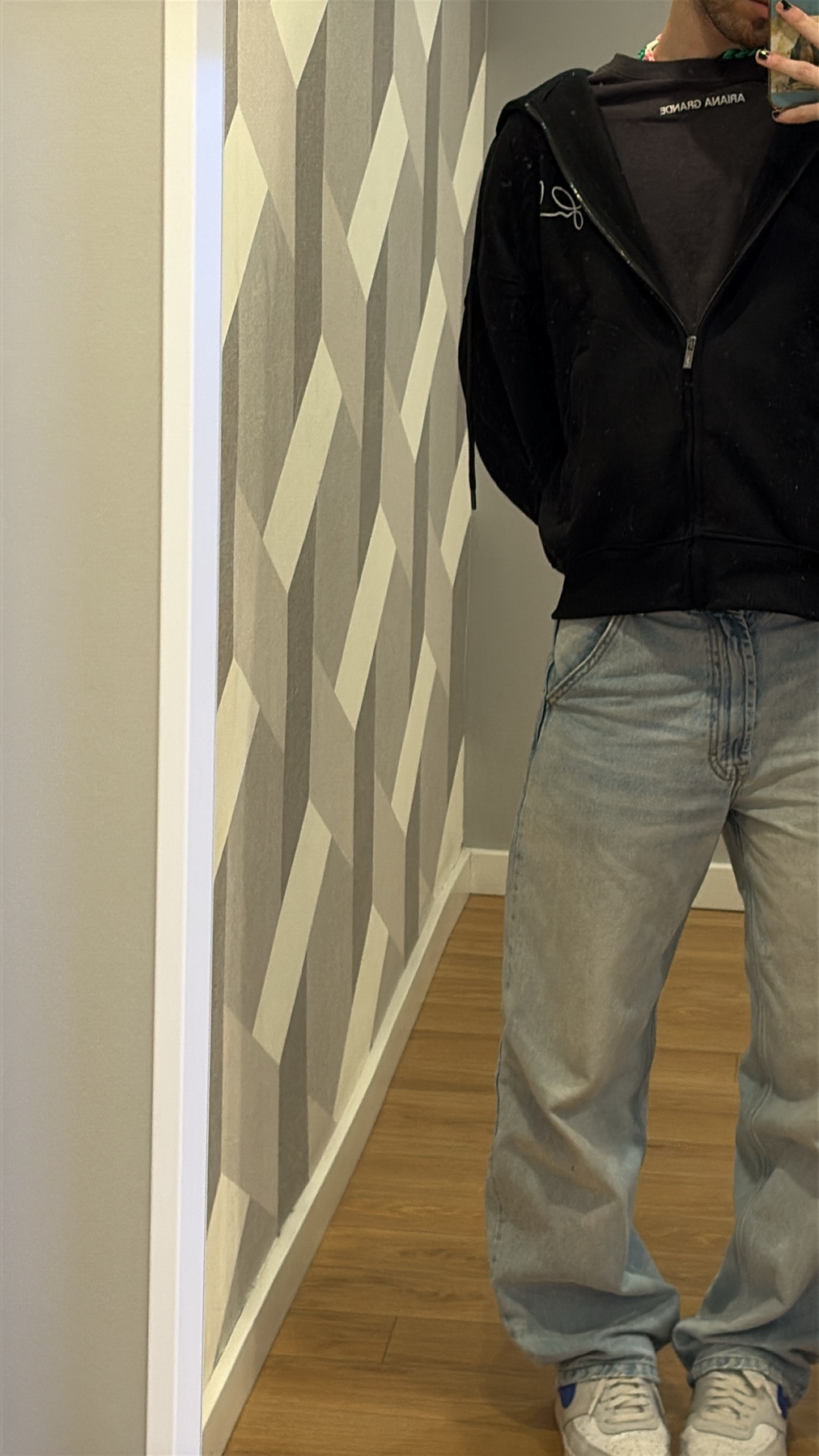 A man poses in a mirror, showcasing his outfit. He wears a black jacket, jeans, and sneakers.