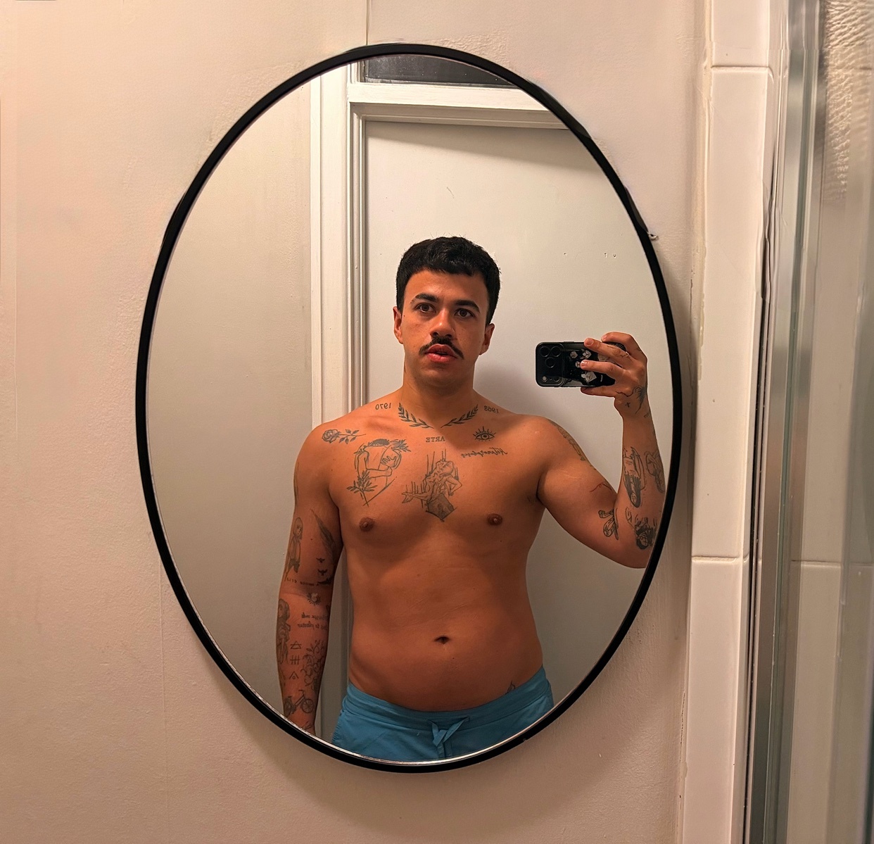 A man with a mustache and tattoos takes a mirror selfie in a bathroom. He is shirtless and wearing blue shorts.