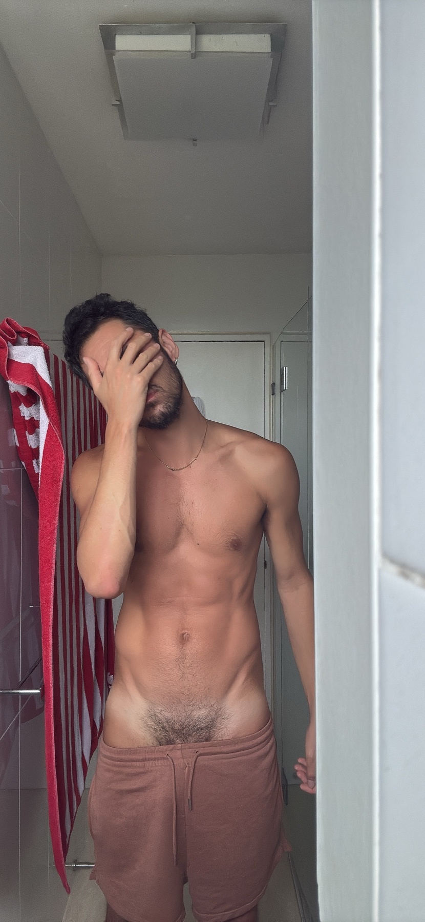 A man with dark hair and stubble stands shirtless in a bathroom, covering his face with his hand. He is wearing brown shorts and a towel hangs on a rack behind him.