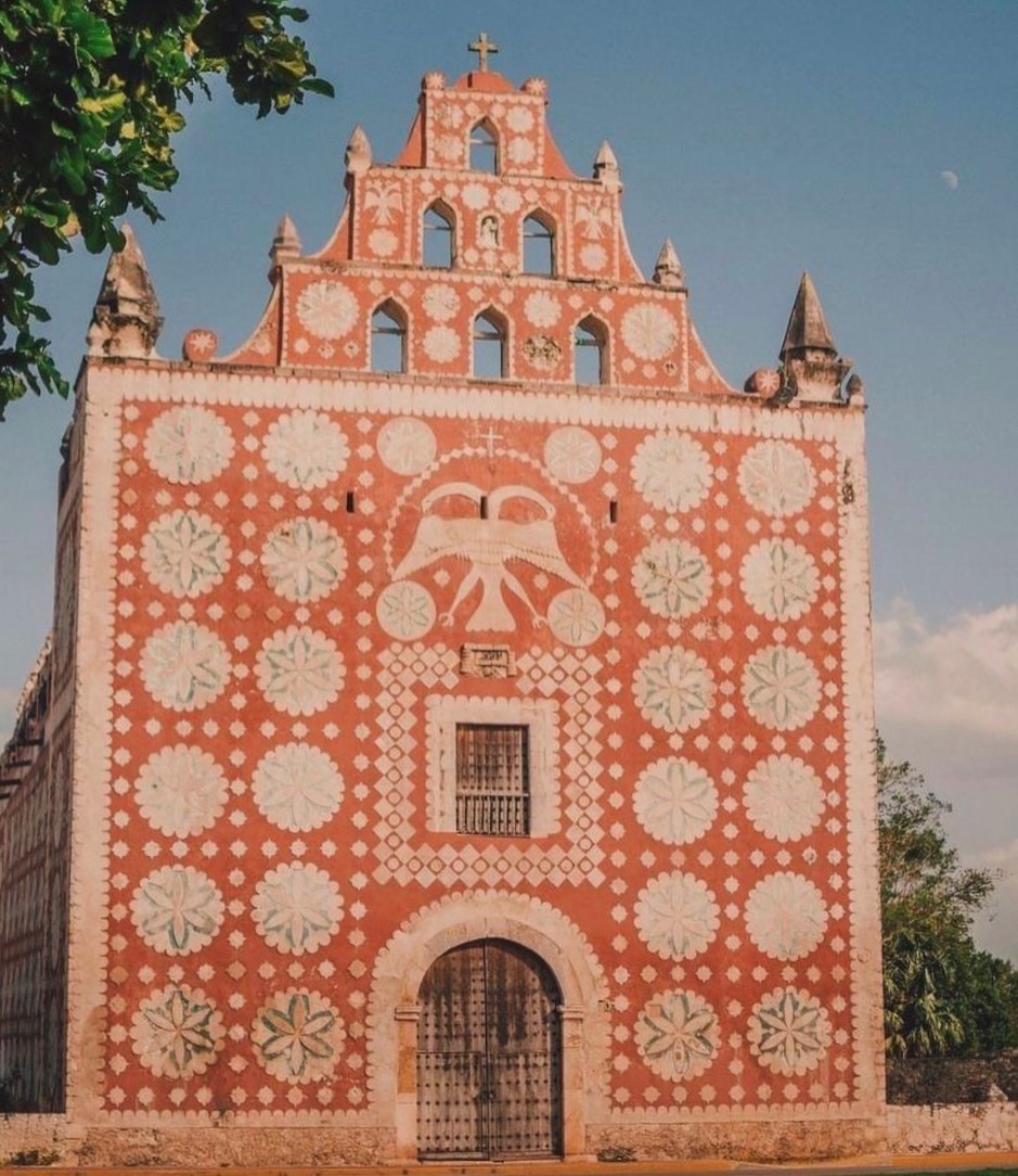 A vibrant, ornate church facade in Mexico features intricate white and blue floral patterns against a striking red background. The building's unique architectural style is highlighted by its decorative elements and arched wooden doorway.
