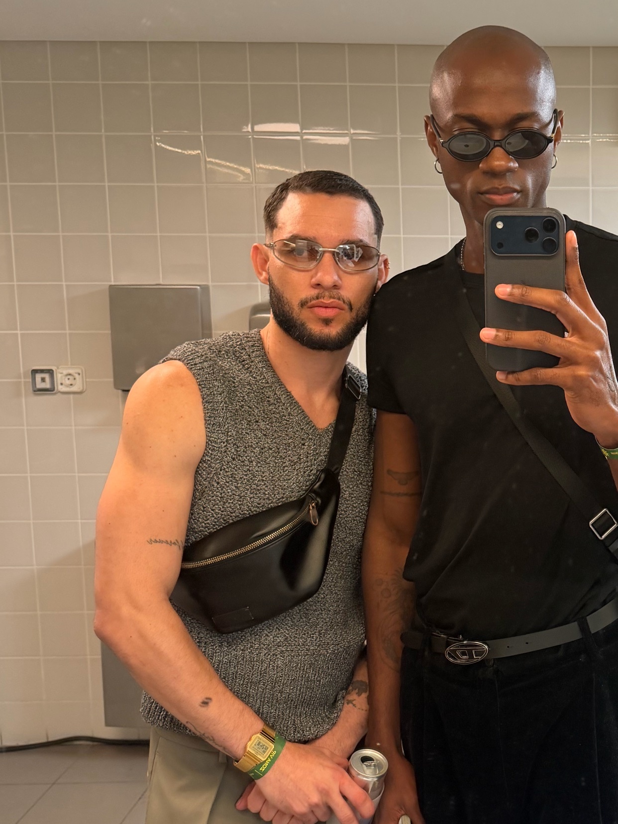 Two men pose for a mirror selfie in a bathroom. They are both wearing stylish outfits and sunglasses.