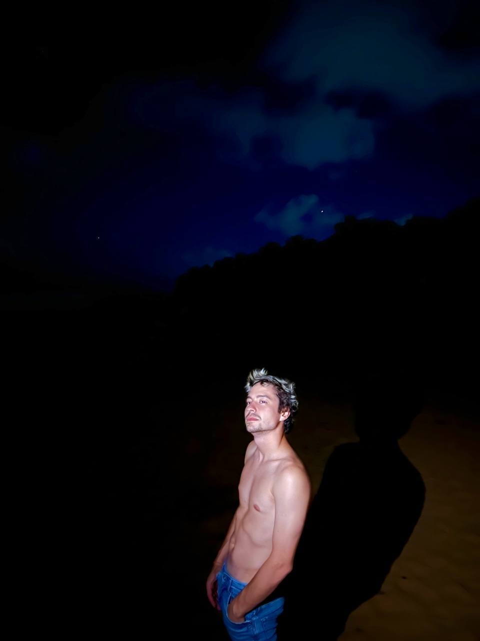 A man stands shirtless on a beach at night, looking up at the sky. The photo has a dark, moody aesthetic.