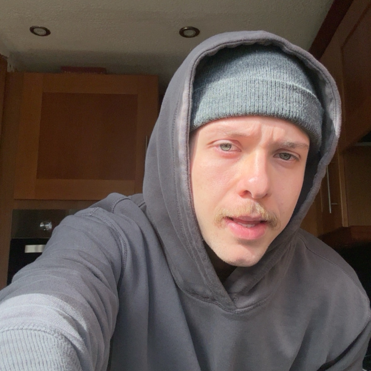 A man with a mustache wears a gray beanie and a dark gray hoodie. He is looking directly at the camera in a selfie-style shot.