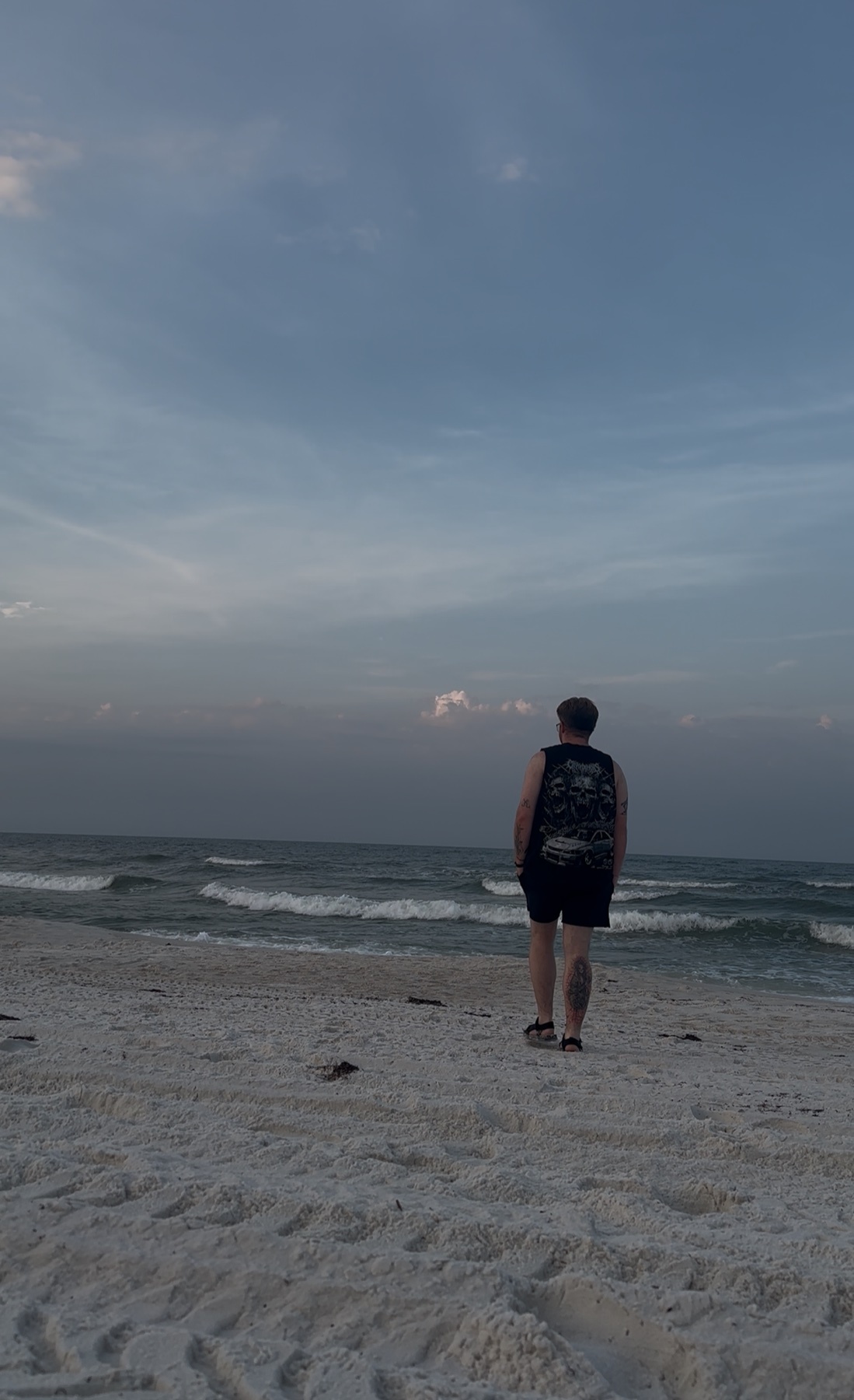 A man stands on a sandy beach, looking out at the ocean waves under a cloudy sky. The image captures a serene moment of solitude by the sea.