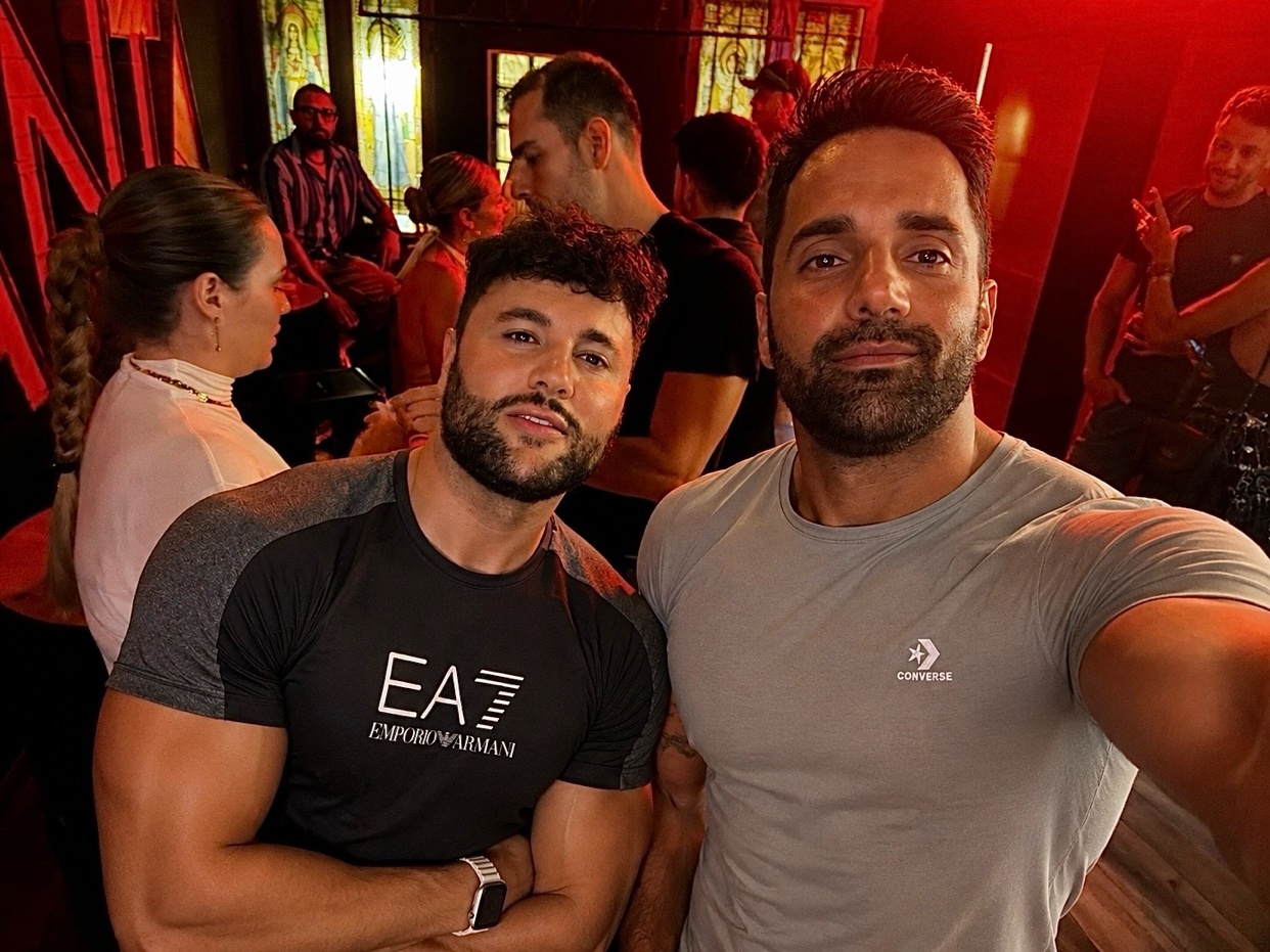 Two men take a selfie in a crowded, dimly lit venue. The man in the foreground on the left wears a black t-shirt with white lettering, while the man on the right wears a grey t-shirt. Both men have beards and appear to be in good spirits.
