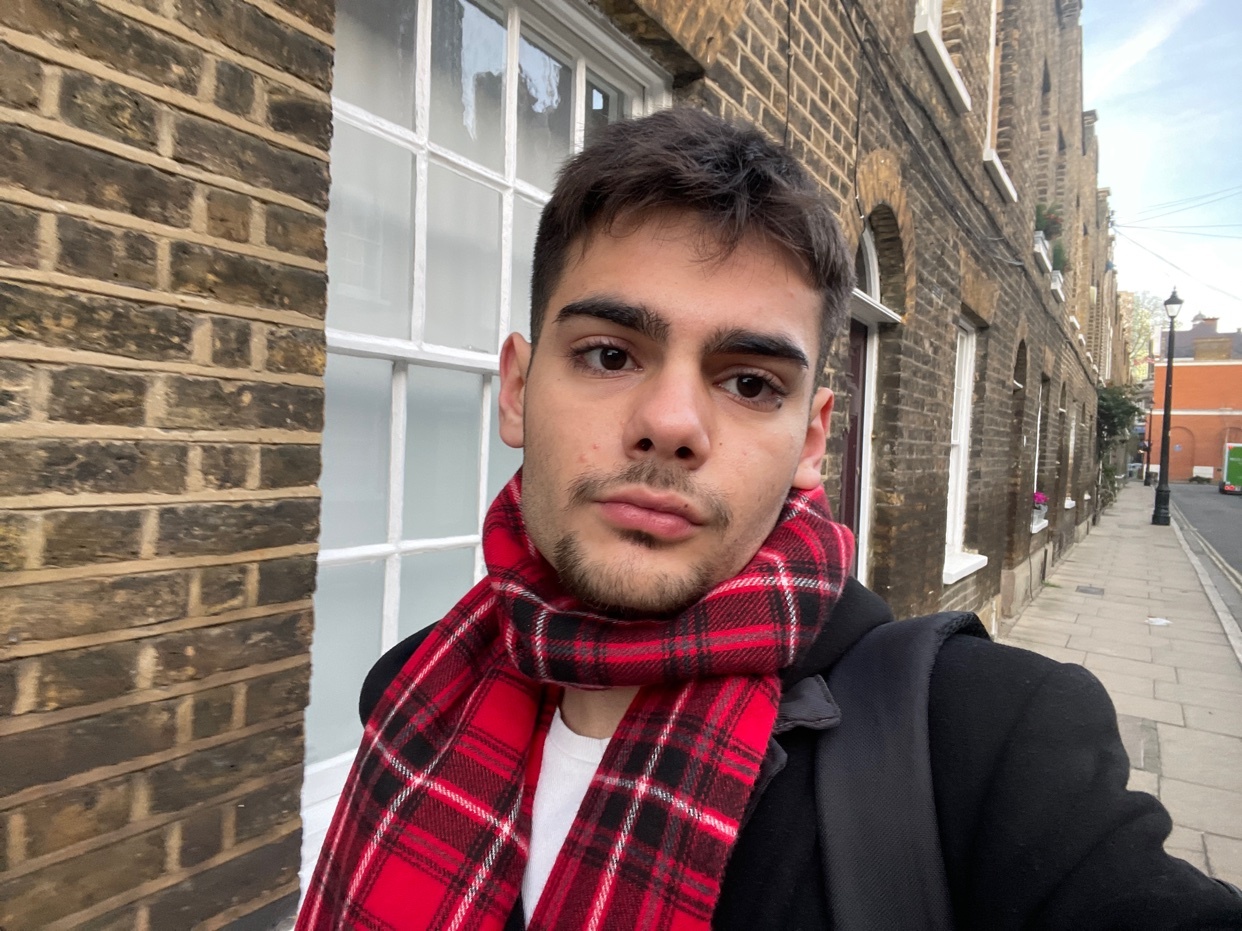 A man poses for a selfie in front of a brick building. He is wearing a red plaid scarf and a black jacket.