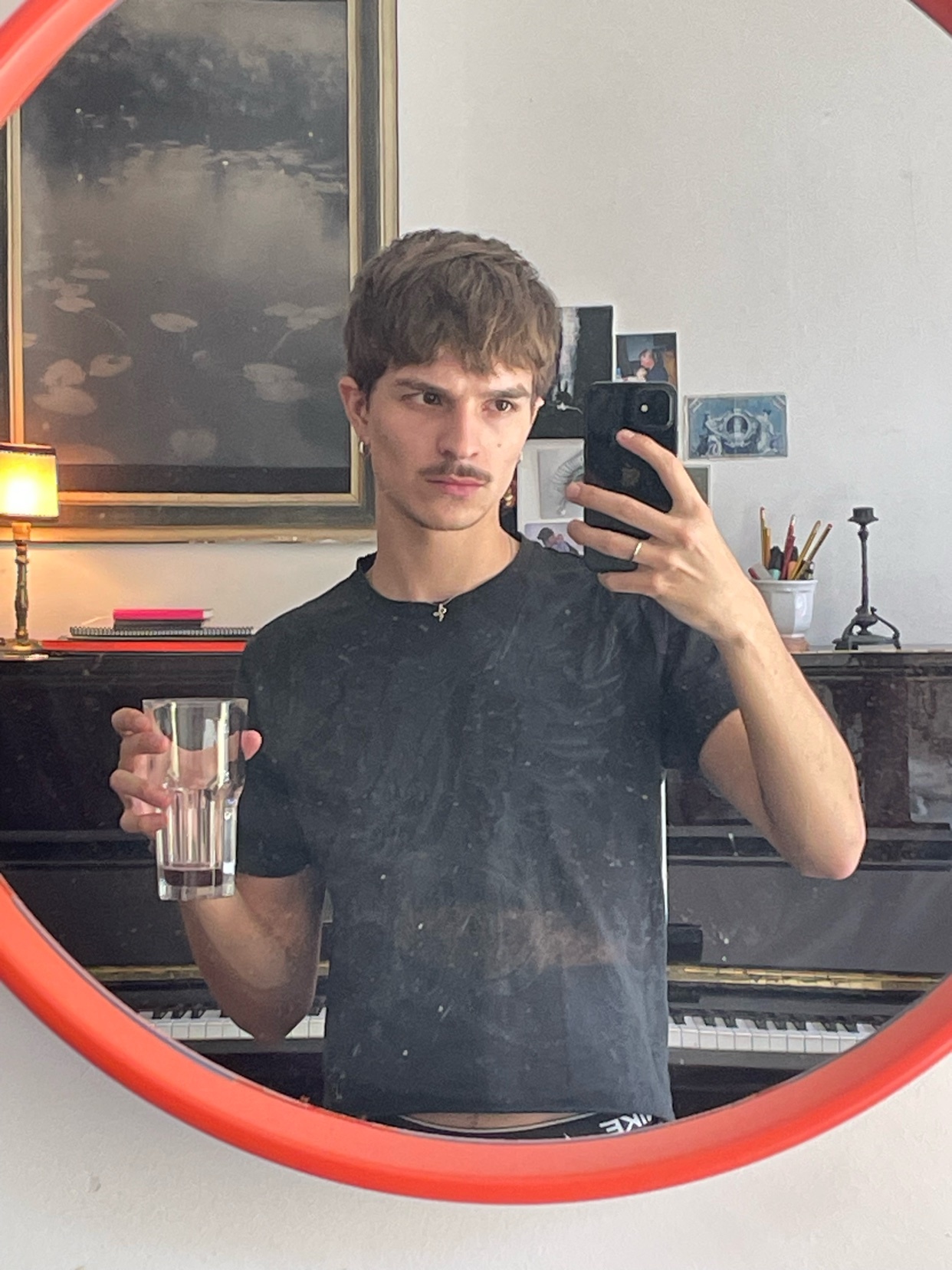 A man takes a mirror selfie, holding a glass of water. The photo is taken in a room with a piano and artwork.