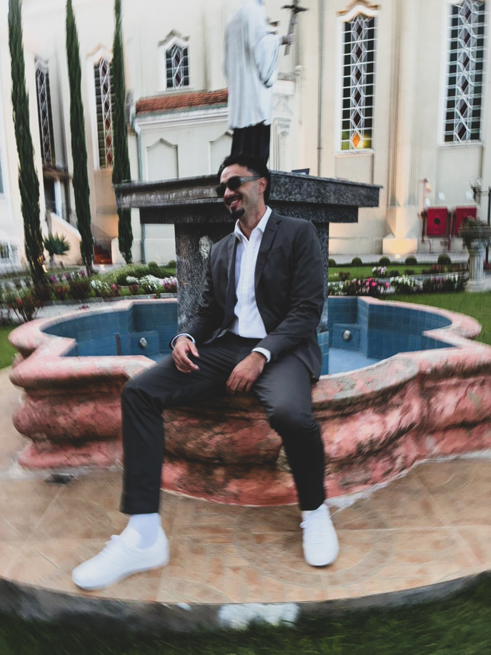 A man in a suit and sunglasses sits on the edge of a fountain in front of a building with ornate windows. The image has a blurred effect, giving it a dynamic feel.