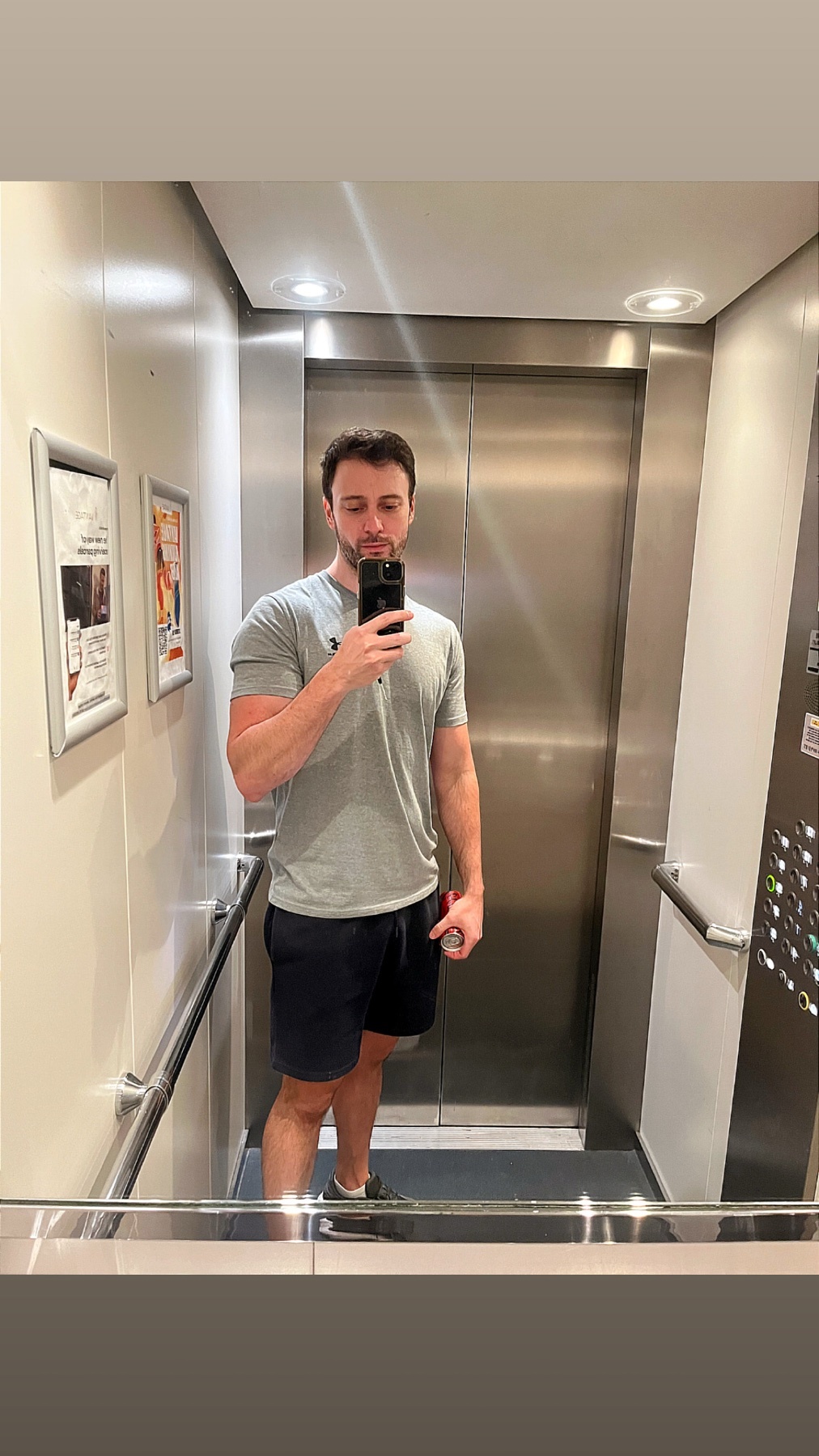 A man takes a mirror selfie in an elevator, wearing a grey t-shirt and dark shorts.