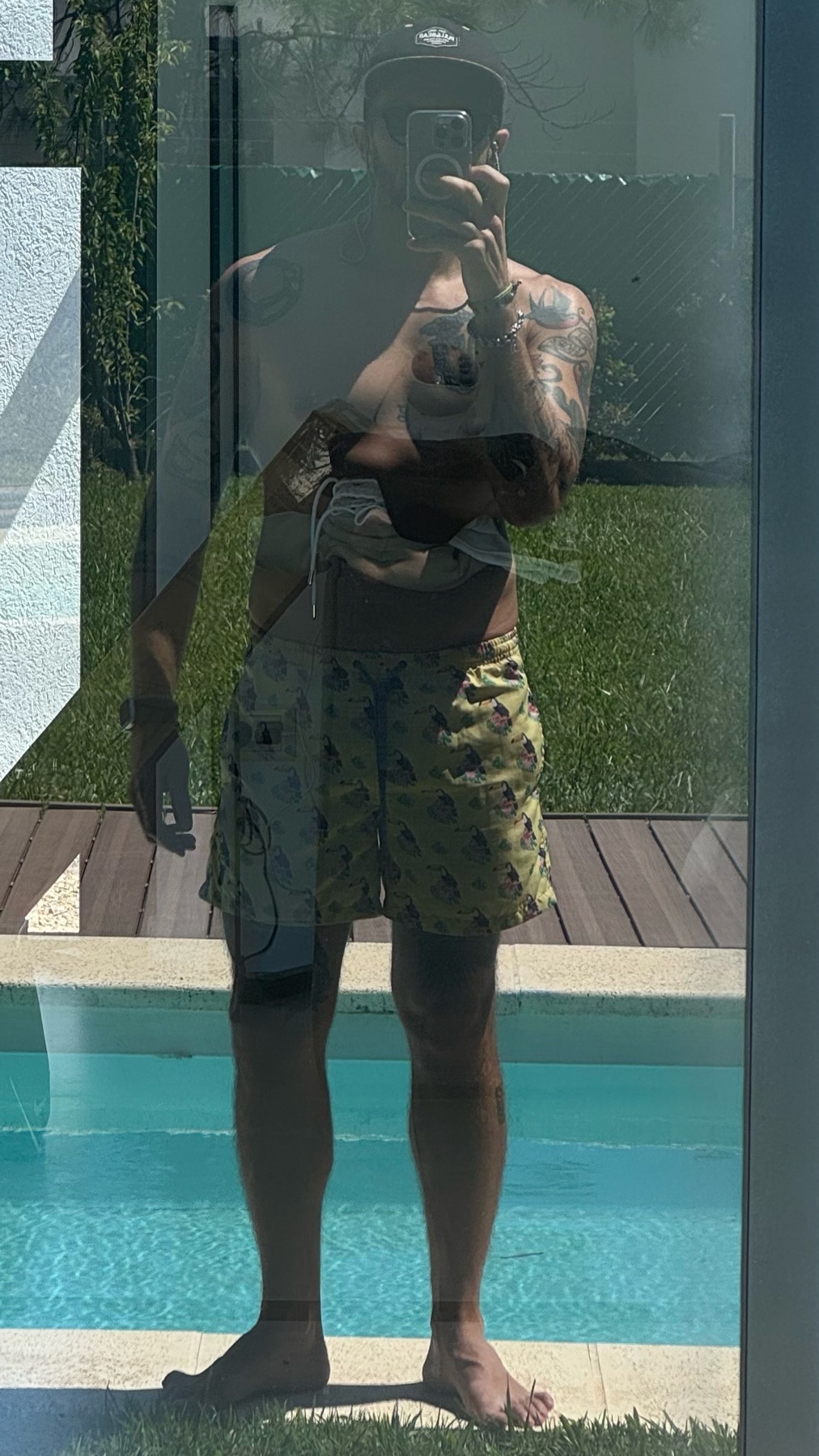 A man takes a mirror selfie in his swimwear. The photo is taken outdoors near a pool.