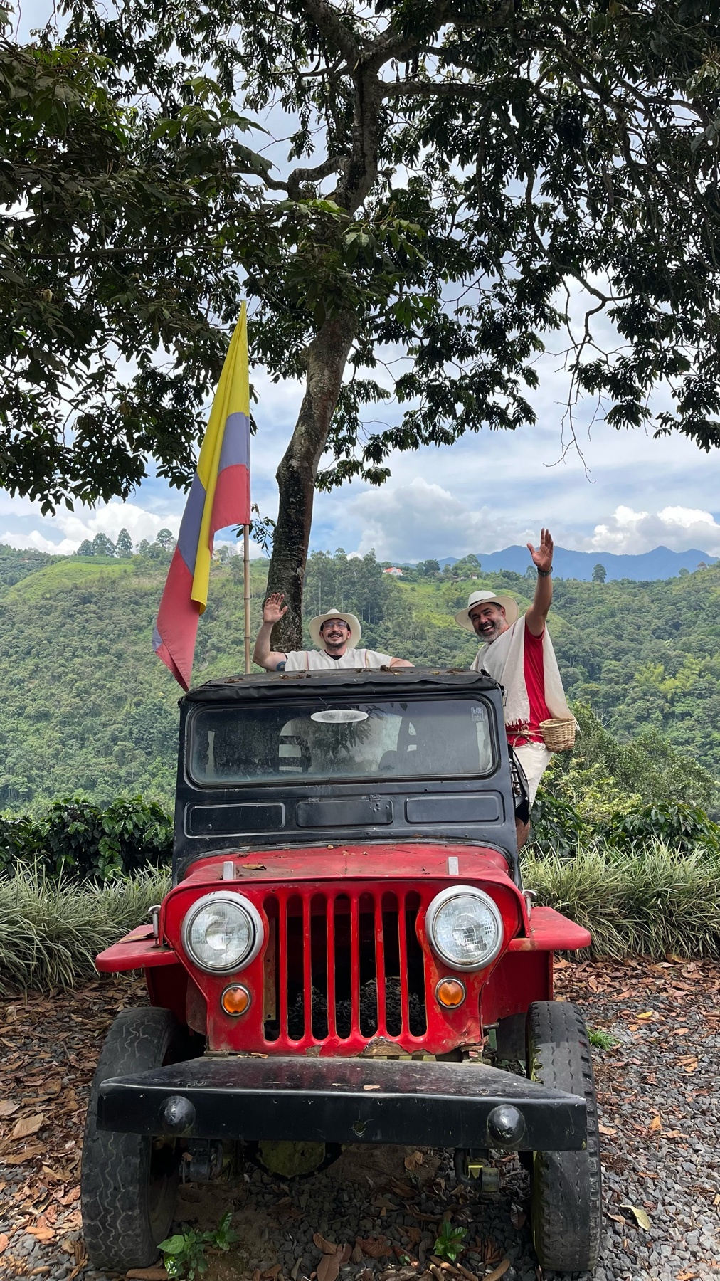Two men wave from a vintage red jeep parked on a coffee farm, with a Colombian flag flying behind them. The lush, green landscape of rolling hills and trees stretches out in the background under a cloudy sky.