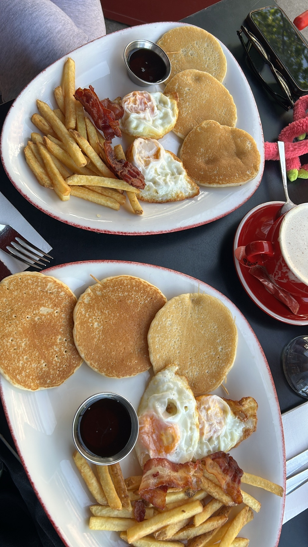 Two plates of breakfast are shown, each featuring pancakes, fried eggs, bacon, and french fries. A cup of coffee sits to the side.