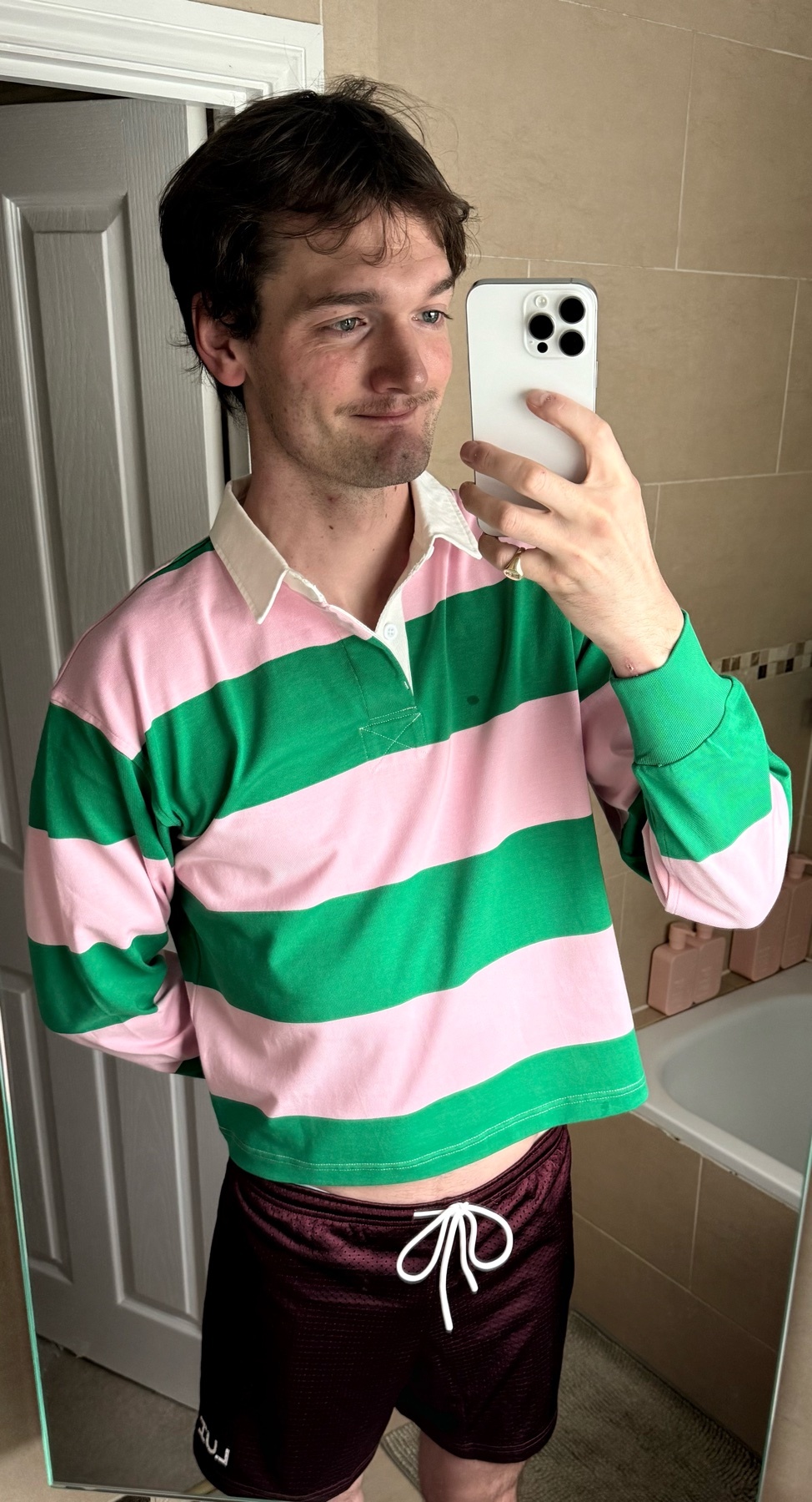 A man takes a mirror selfie in a bathroom. He is wearing a striped shirt and shorts.