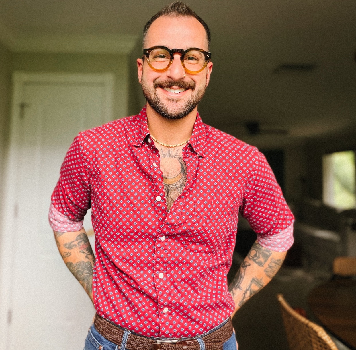 A man with short brown hair, a beard, and glasses smiles at the camera. He is wearing a red patterned button-down shirt and blue jeans, with his hands on his hips.