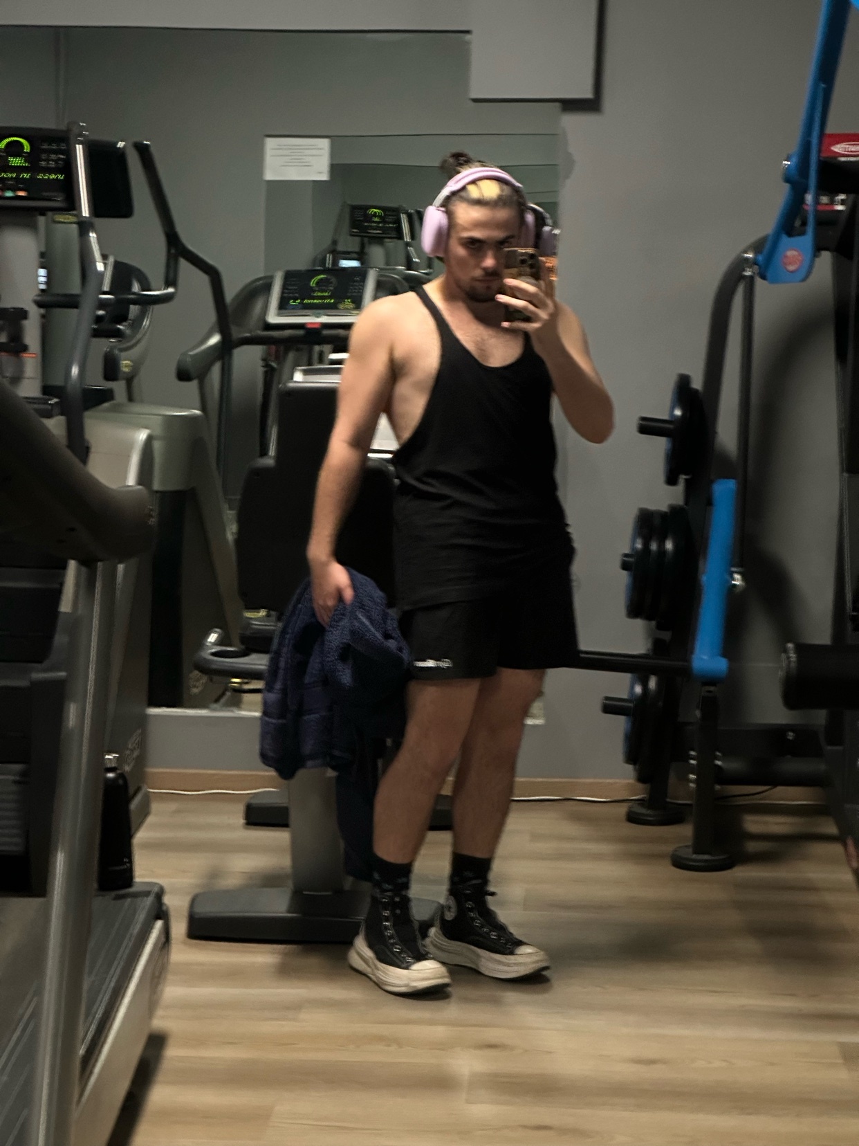 A man takes a mirror selfie at the gym. He is wearing a black tank top and shorts.