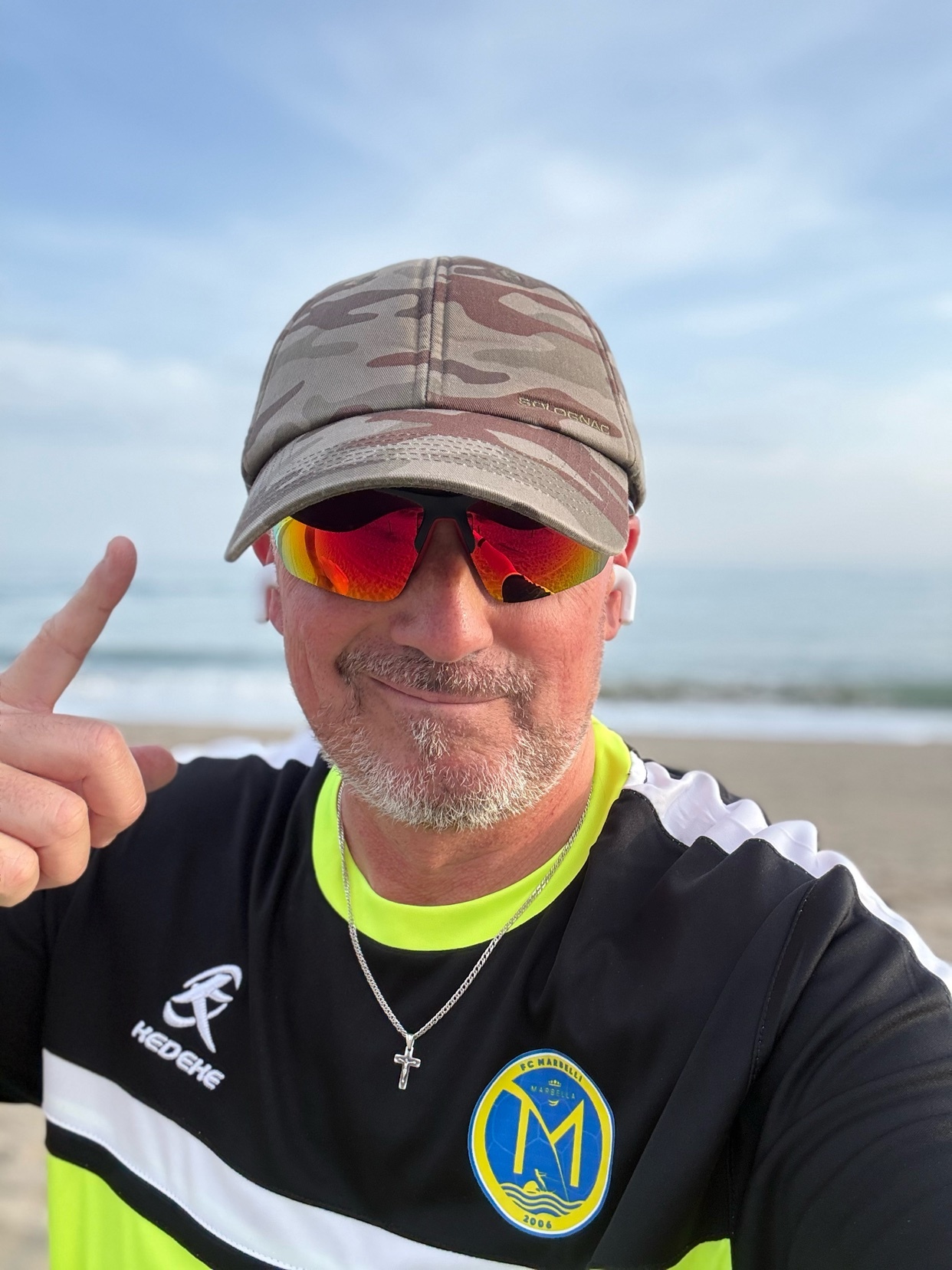 A man wearing sunglasses and a baseball cap poses for a selfie on the beach. He is wearing a black and neon yellow athletic shirt and a silver cross necklace.