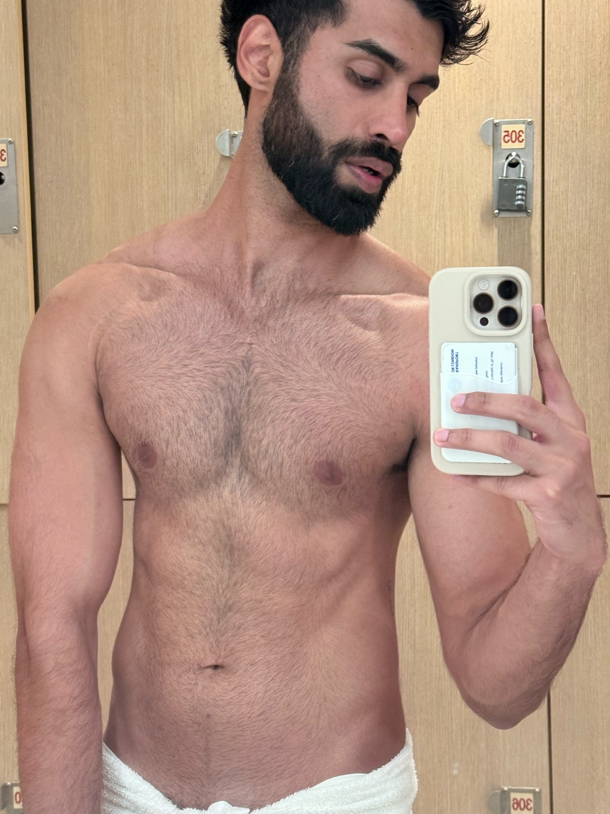 A man with a beard and dark hair takes a selfie in a locker room, wearing a towel around his waist.