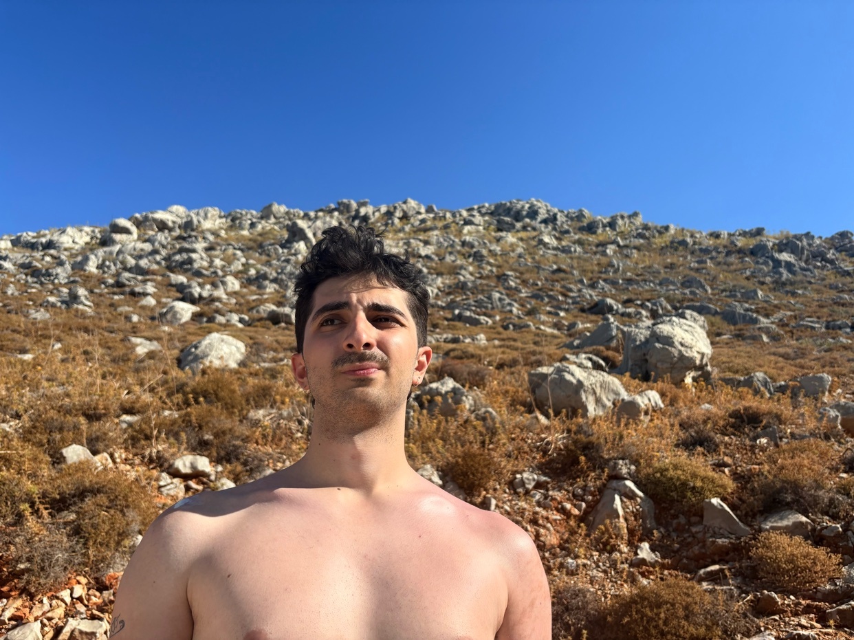 A man stands shirtless in front of a mountain. The sky is blue and the sun is shining.