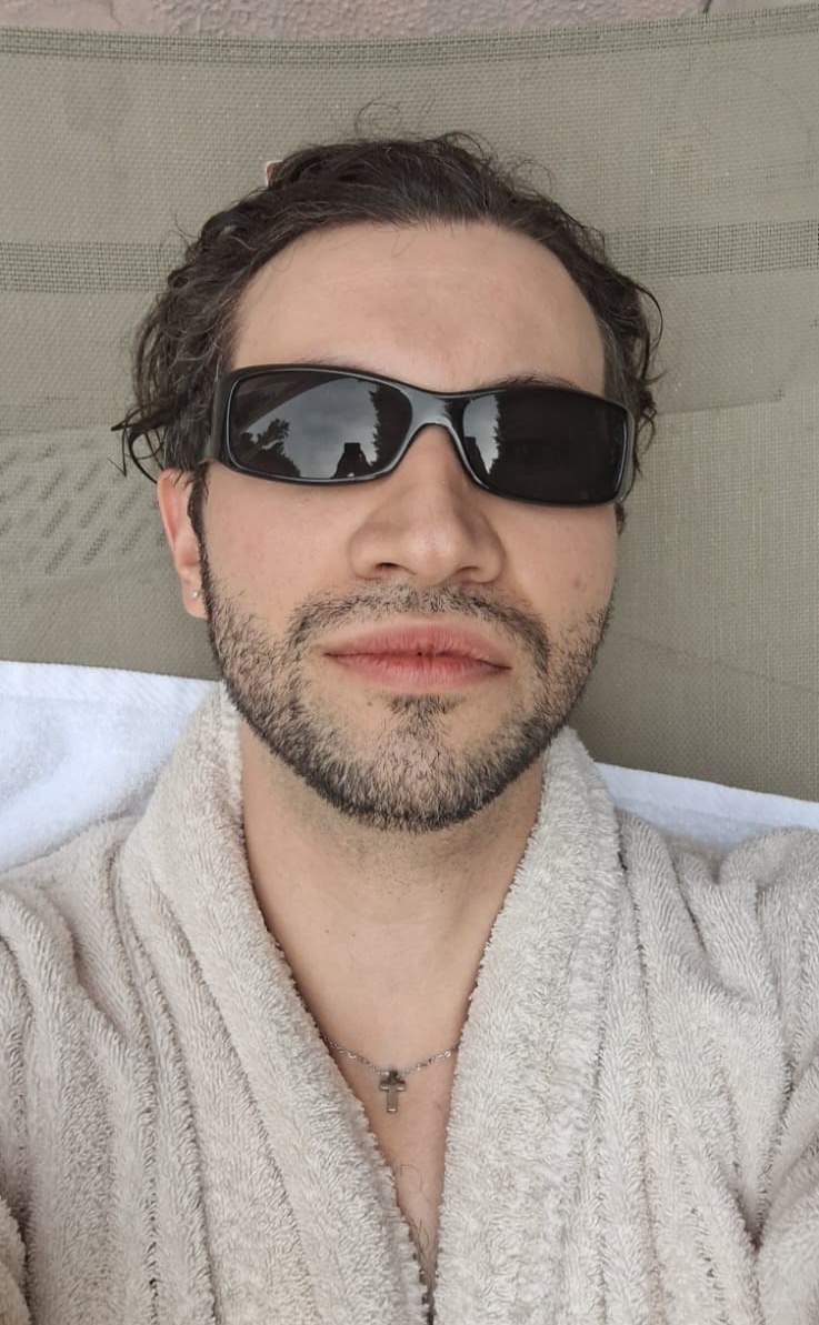 A man wearing sunglasses and a robe poses for a selfie, looking relaxed.